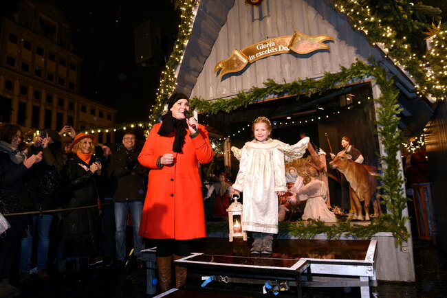 Vor dem Hintergrund einer lebensgroßen Weihnachtskrippe stehen eine Frau in rotem Mantel und ein als Engel gekleidetes blondes Kind, das eine Laterne hält.