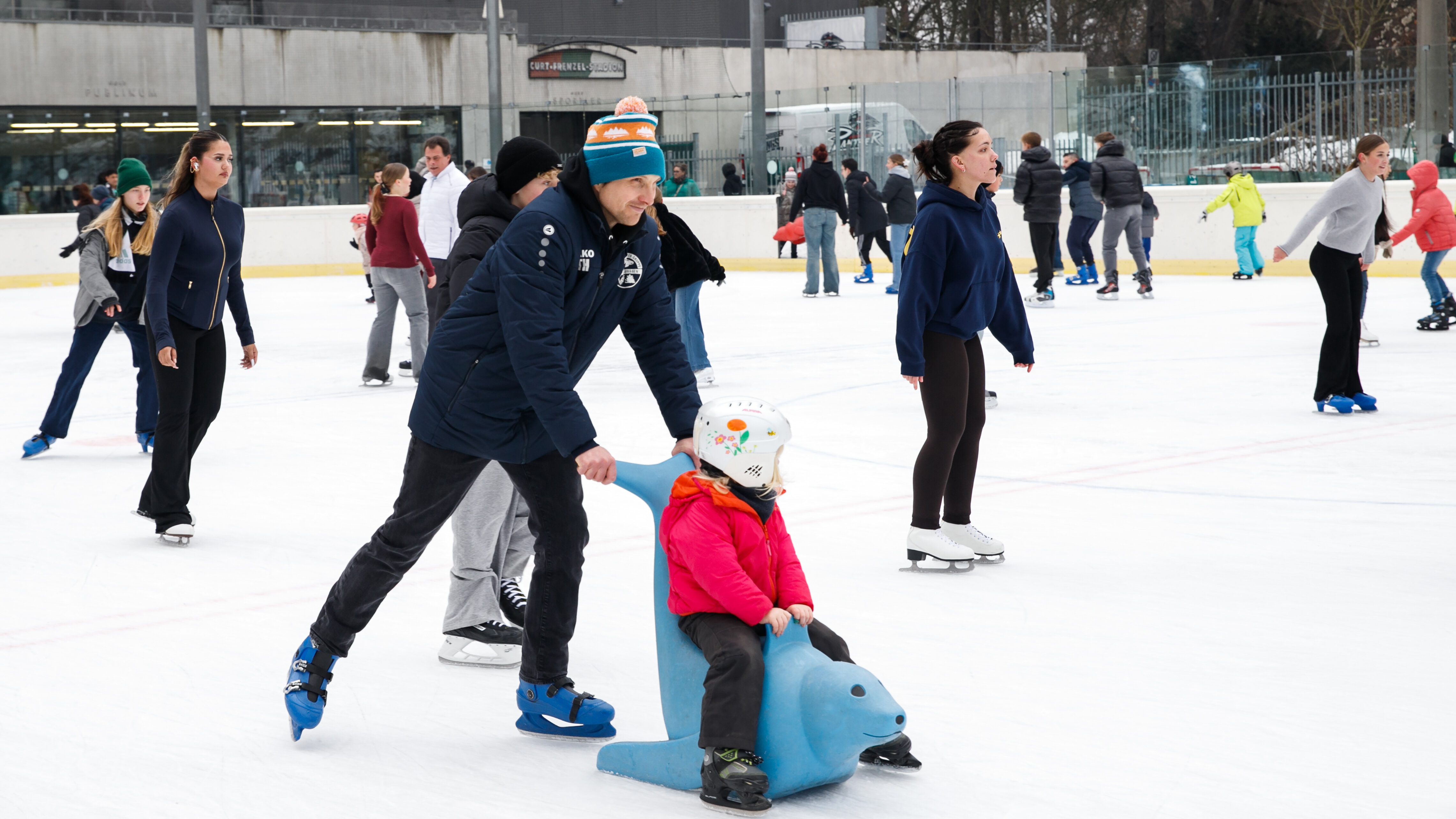 Das Foto zeigt einen Eisfläche auf der viele Leute Schlittschuh laufen. In der Mitte vom Bild ist ein Mann und ein Kind. Das Kind sitzt auf einer Eislaufhilfe in Robbenform, die der Vater anschiebt.