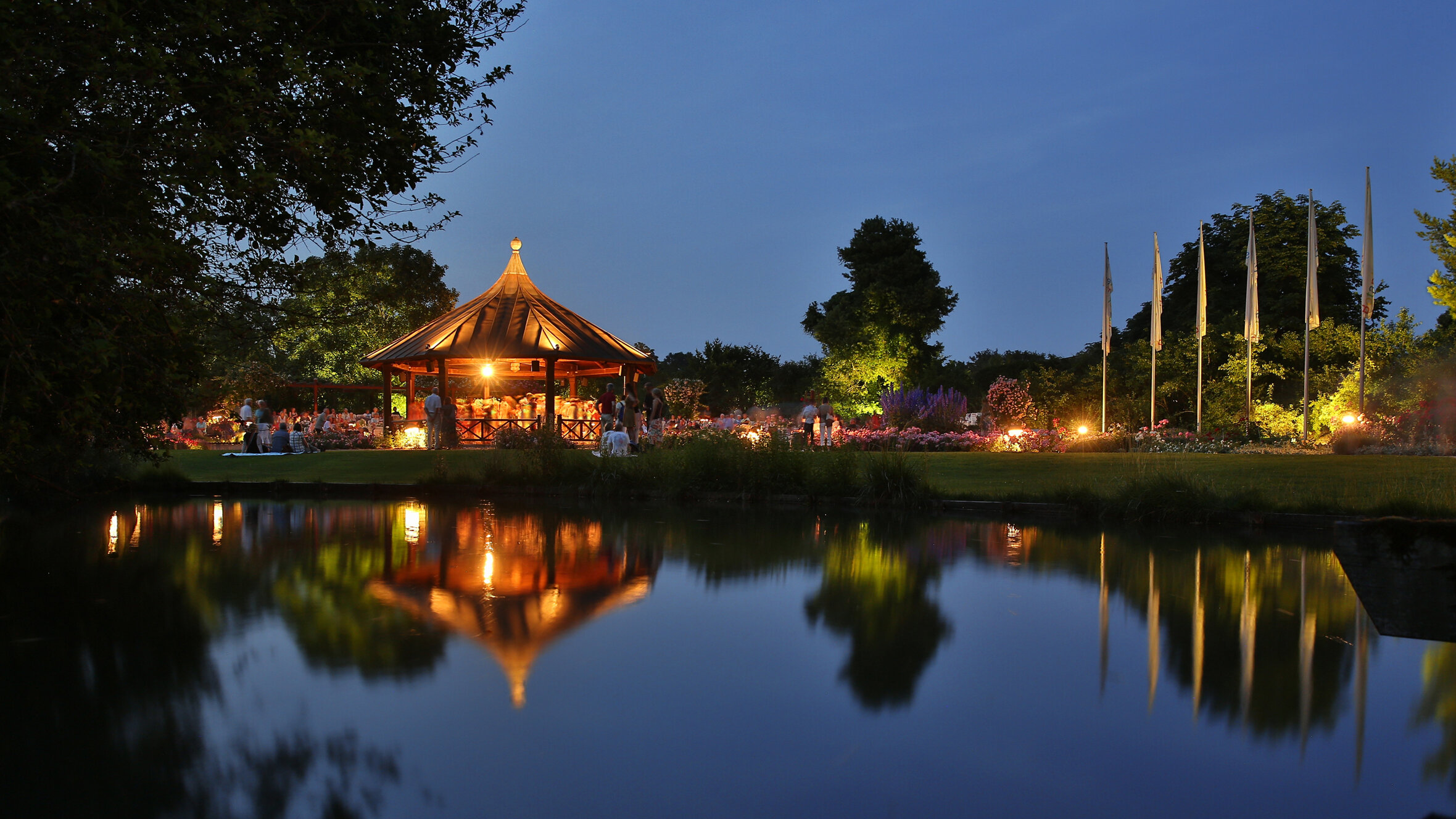 Ein Pavillon im Botanischen Garten ist beleuchtet. Es ist Abend, der Himmel ist dunkelblau. Der Pavillon und die umliegenden Bäume spiegeln sich in einem kleinen See