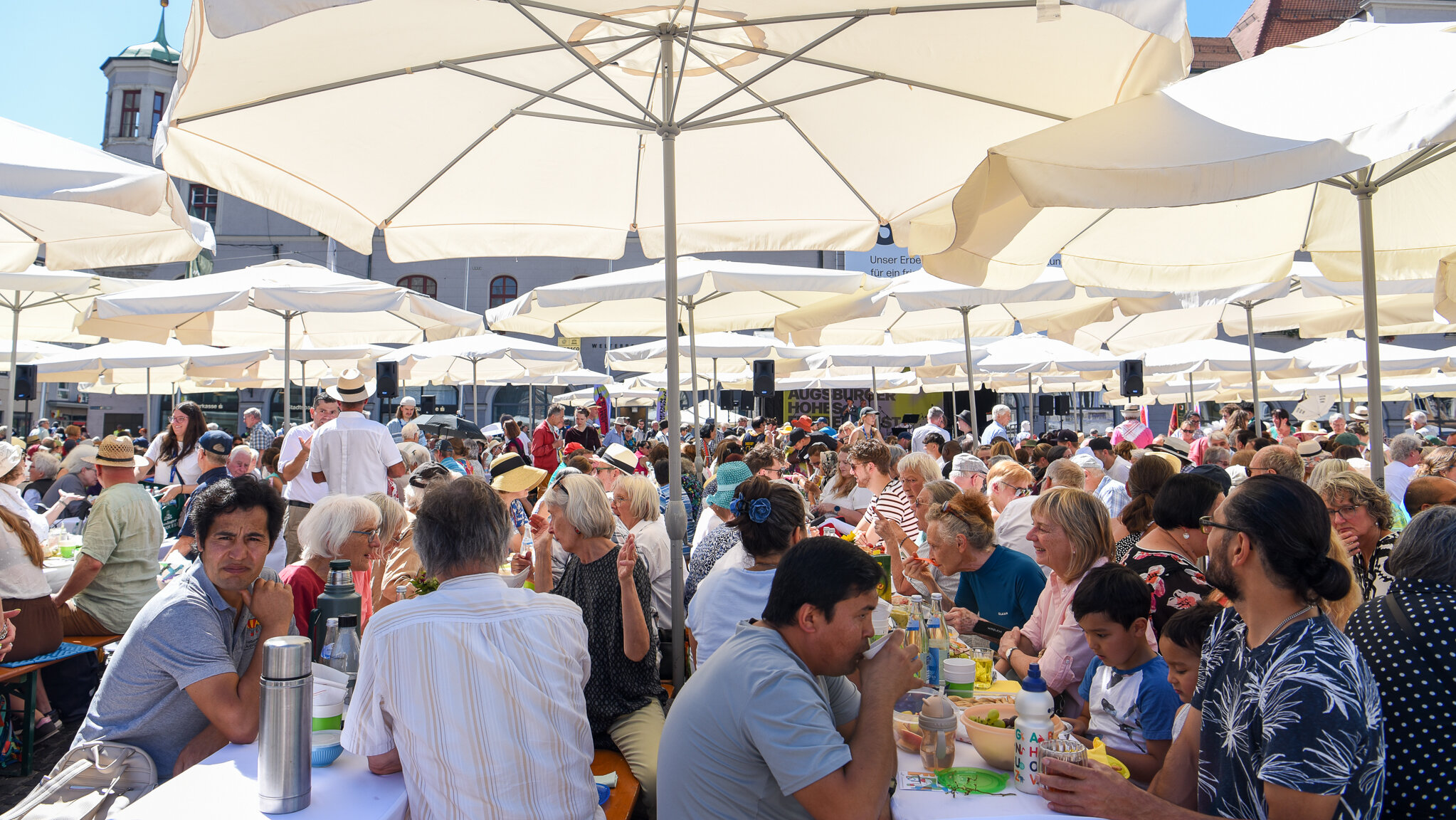 viele Menschen sitzen auf Bierzeltbänken unter freiem Himmel, mit Schirmen vor der Sonne geschützt