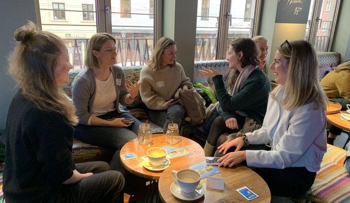 5 Frauen sitzen um einen kleinen Tisch in einem Café und unterhalten sich angeregt