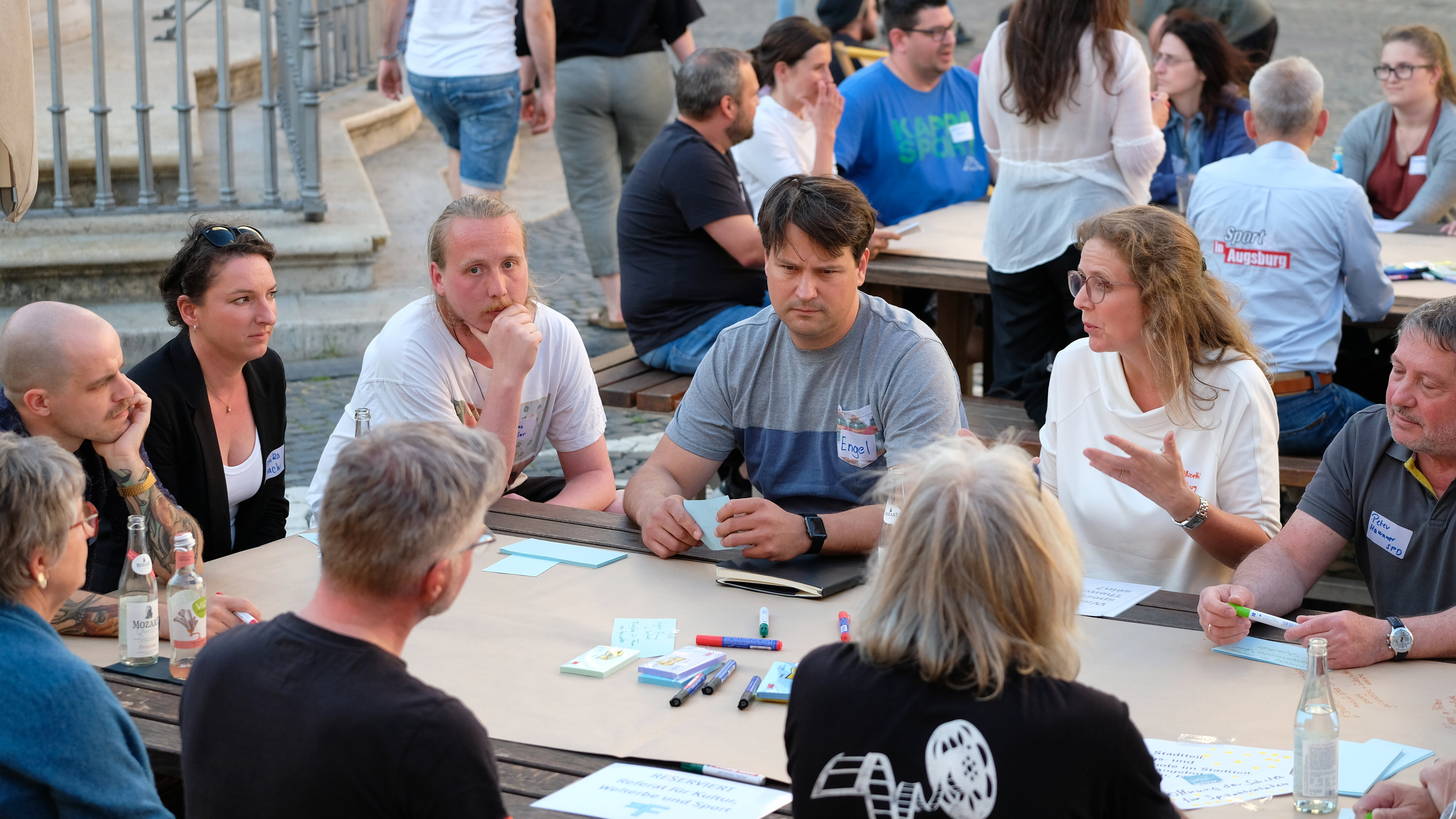 verschiedenste Menschen sitzen auf einem offenen Platz um Tische herum, auf denen Stifte und Papier liegen; sie sind ins Gespräch vertieft.