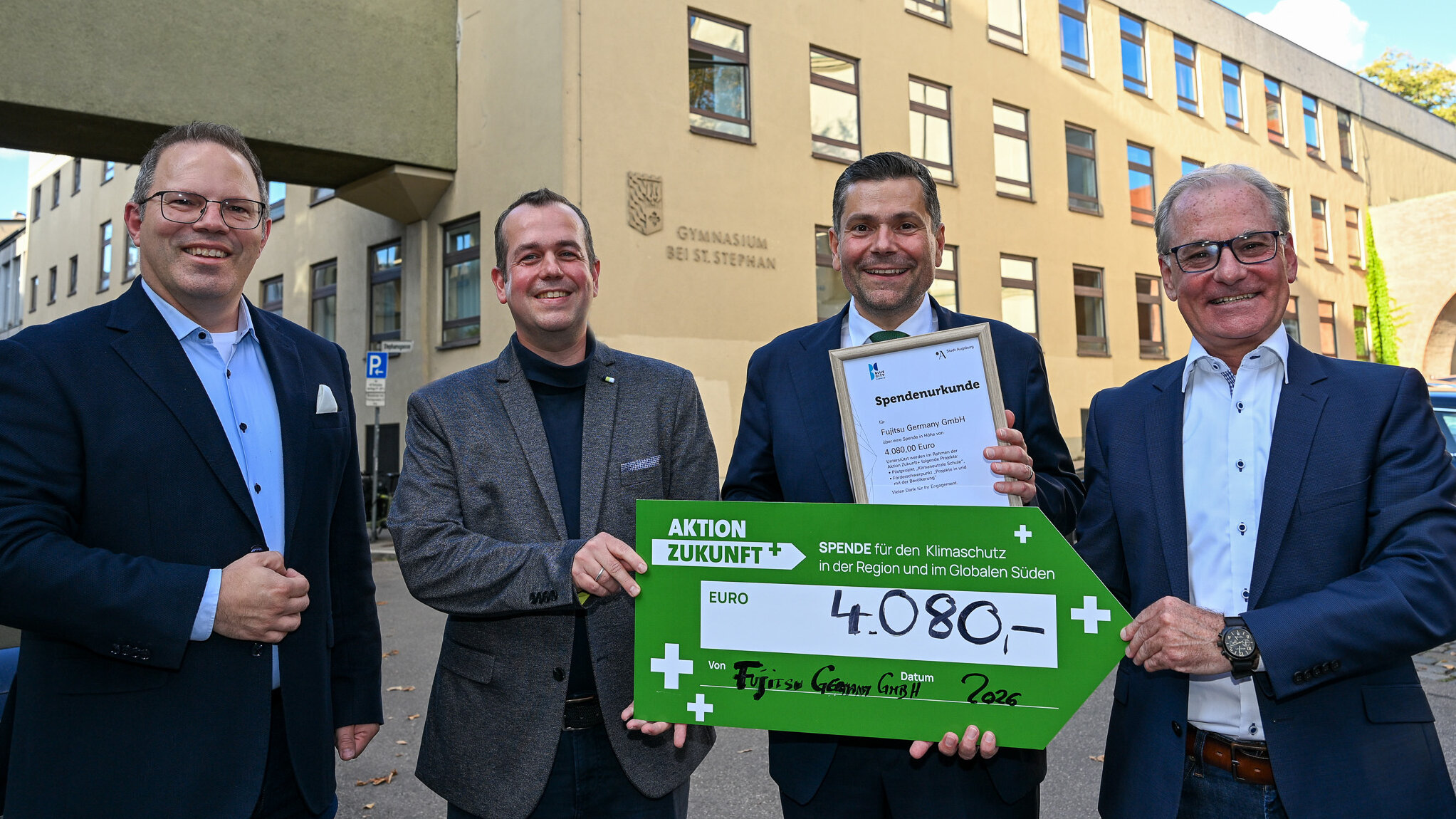 Vier Männer mit einem sehr großen Gutschein vor einem Schulgebäude