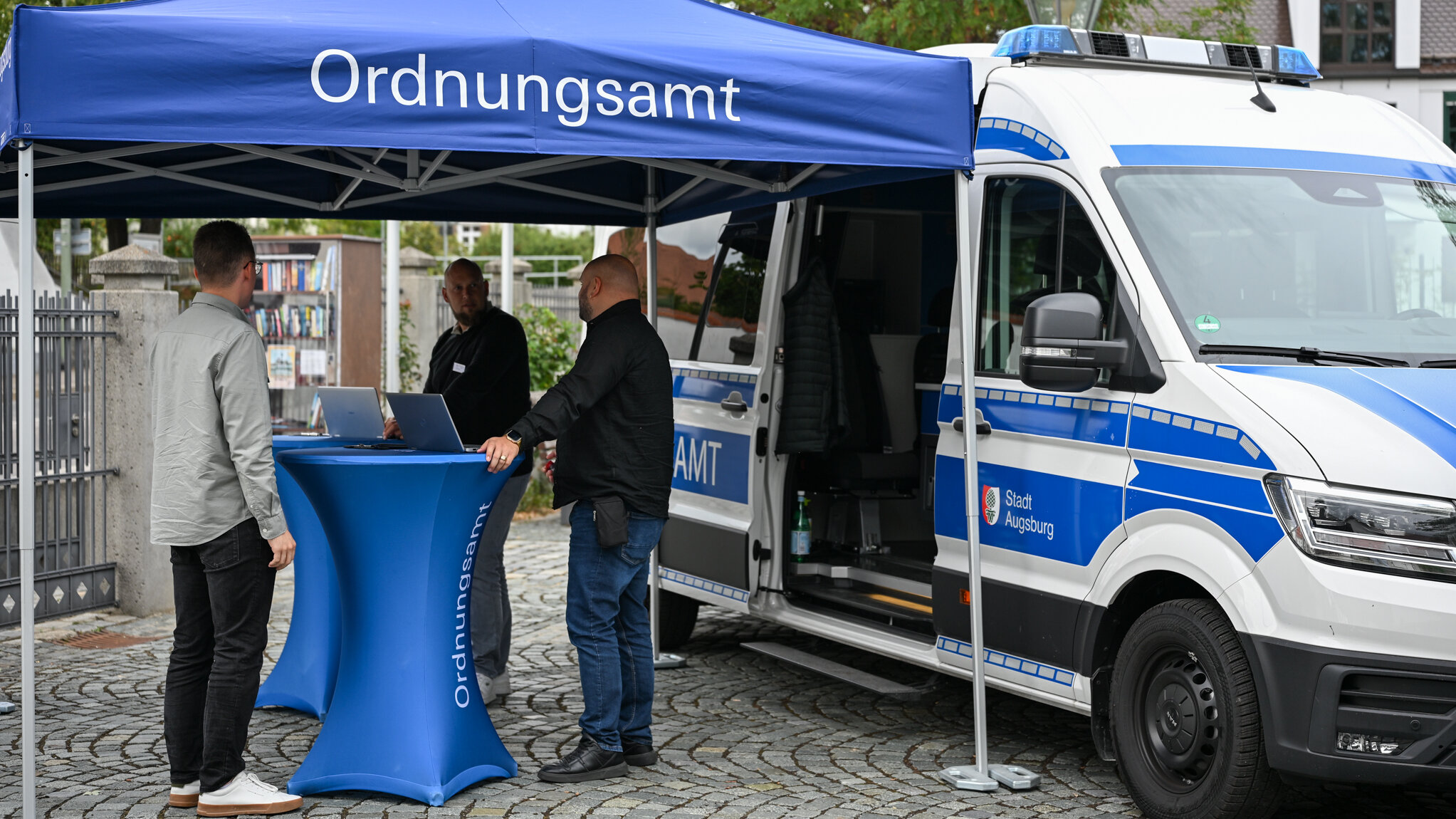 Ein Kastenwagen steht hinter einem Pavillon, unter dem 3 Männer im Gespräch an einem Stehtisch stehen. Zwei von ihnen tragen Uniform.
