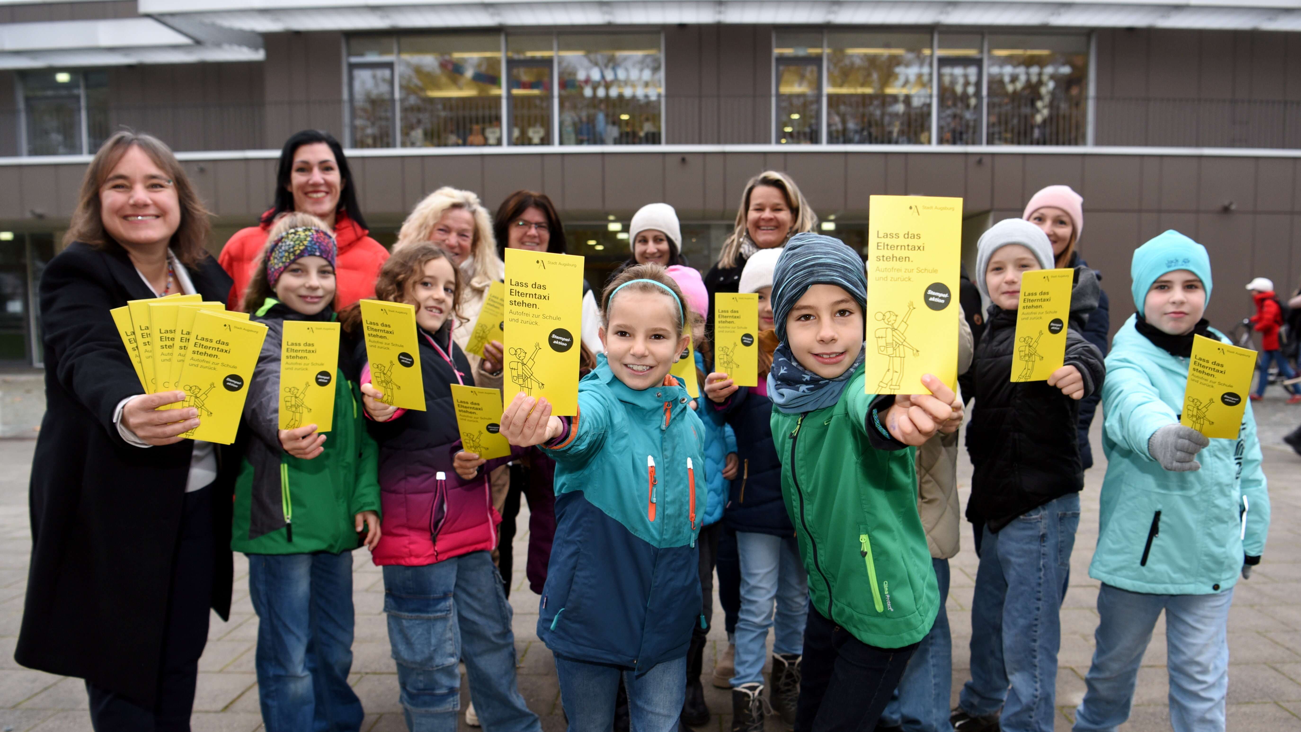 Etwa 10 Kinder und ein paar Erwachsene halten gelbe Zettel in der Hand mit der Aufschrift "Lass das Elterntaxi stehen"