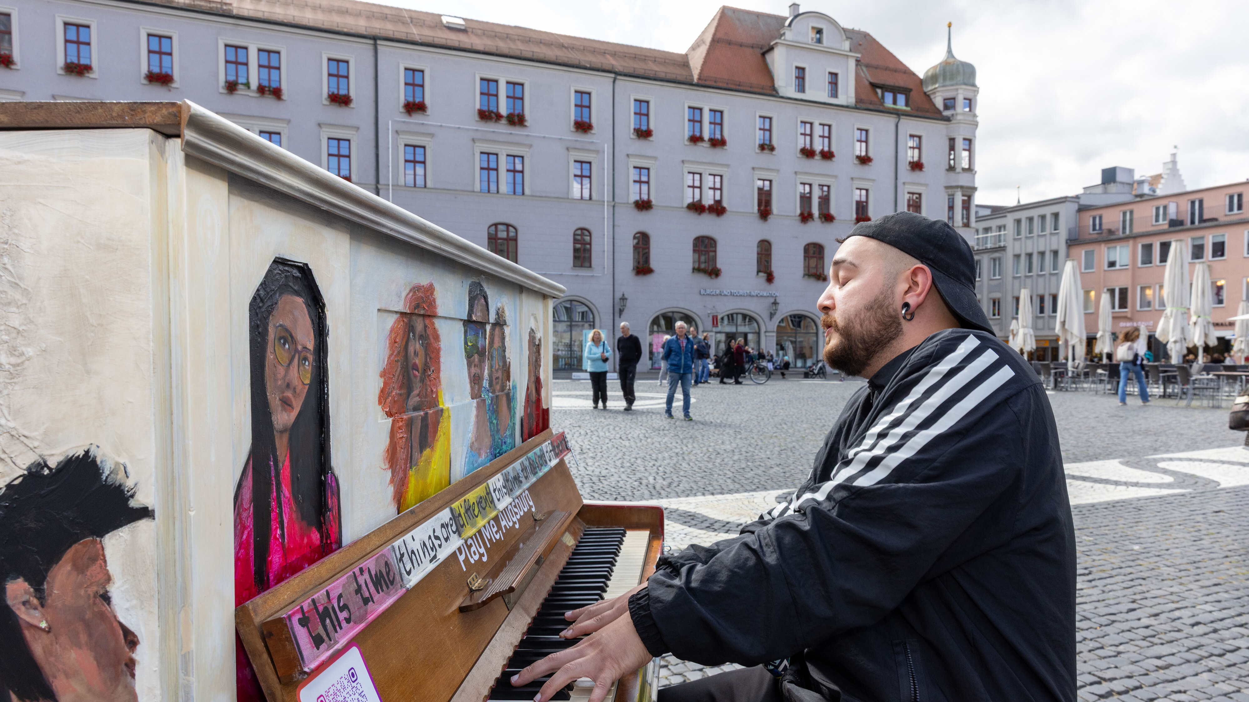 Bemaltes Klavier steht auf dem Augsburger Rathausplatz, ein Mann Mitte 30 sitzt und spielt