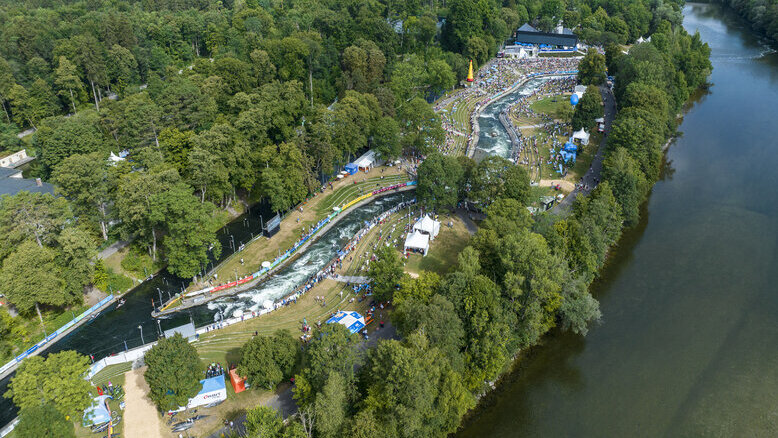 die Kanuslalomstrecke am Augsburger Eiskanal  von oben.  Viele Besuchende jubenl den Kanuten zu. Man kann die Kanustrecke vonm Start bis ins Ziel sehen. Darüber scheint die Sonne. 