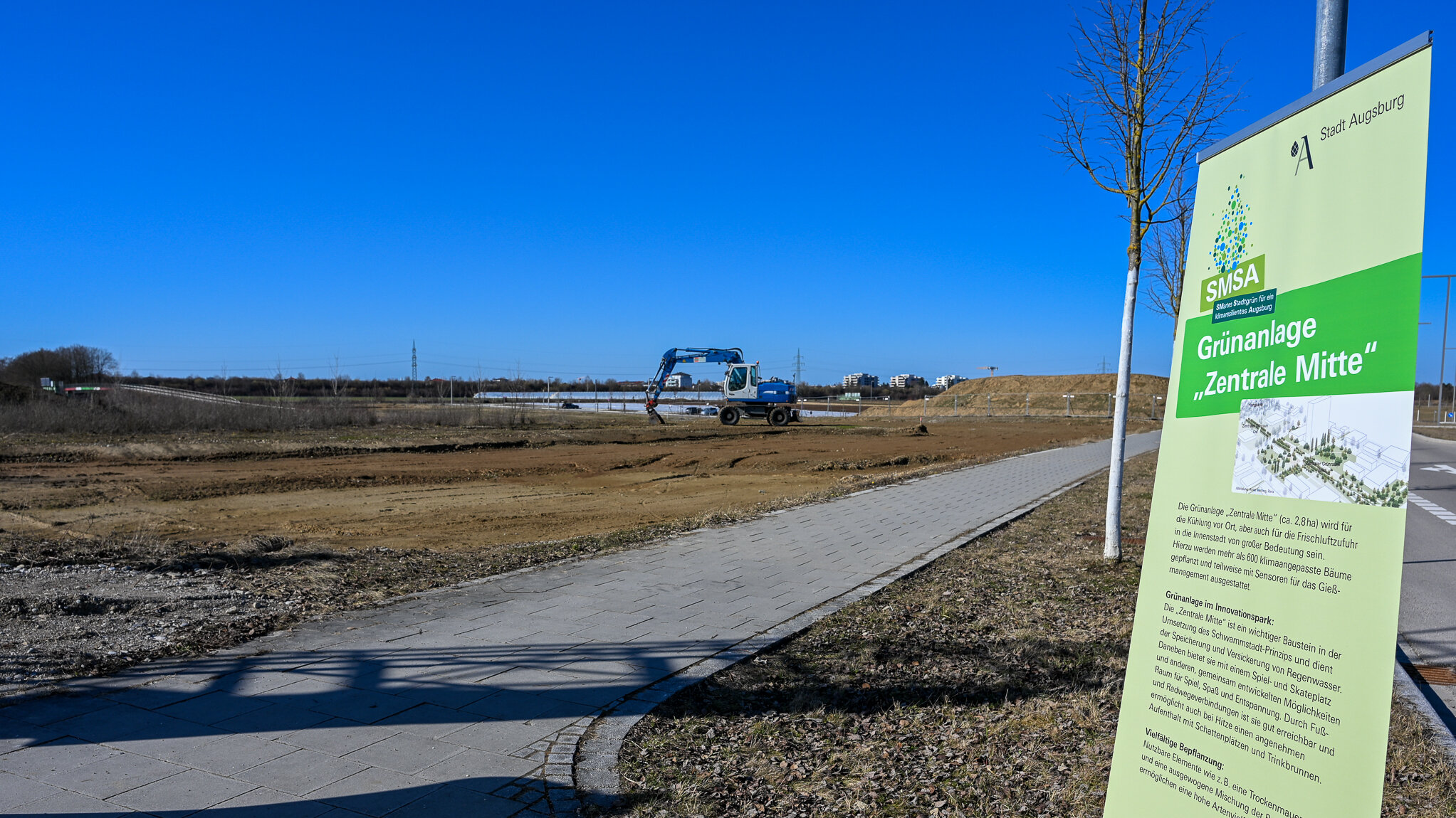 Das Foto zeigt kahle Erdfläche und blauen himmel. Im hintergrund des Bildes ist ein Bagger zu sehen, der in der Erde gräbt. Am rechten Bildrand ist ein großes Plakat auf dem Grünanlage zentrale Mitte steht.