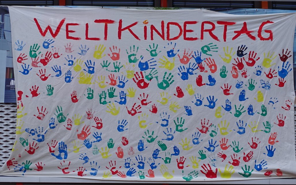 Ein Stoffbanner auf dem in rot Weltkindertag steht. Darunter sind viele bunte Handabrücke.