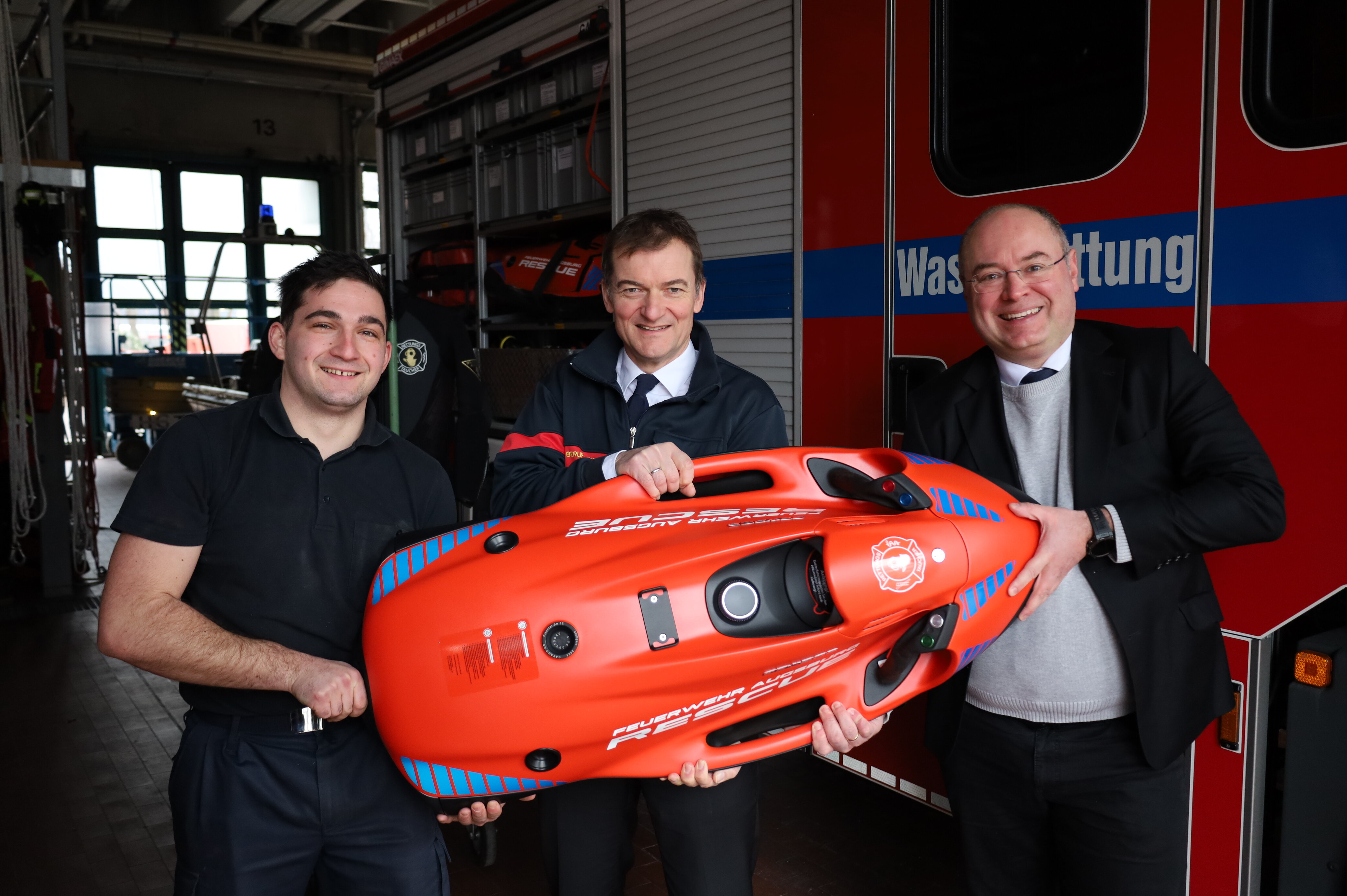 Drei Männer halten einen roten Bob in den Händen. Im Hintergrund steht ein Feuerwehr-Fahrzeug