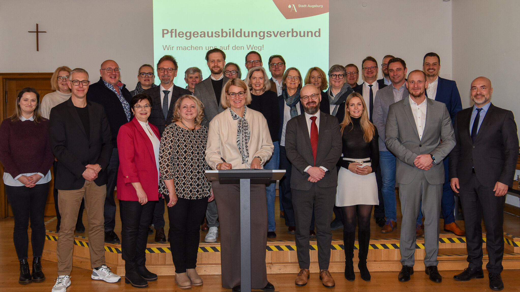 Viele Menschen stehen auf einer Bühne. In der Mitte vorne steht ein Rednerpult auf dem ein Buch liegt.