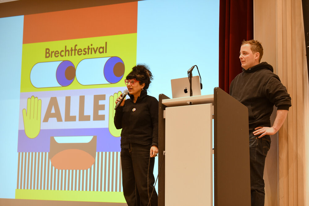 ein Mann und eine Frau, beide jünger, stehen am Rednerpult, im Hintergrund das Logo des diesjährigen Brechtfestivals mit dem Schriftzug "Alle"
