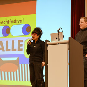 ein Mann und eine Frau, beide jünger, stehen am Rednerpult, im Hintergrund das Logo des diesjährigen Brechtfestivals mit dem Schriftzug "Alle"