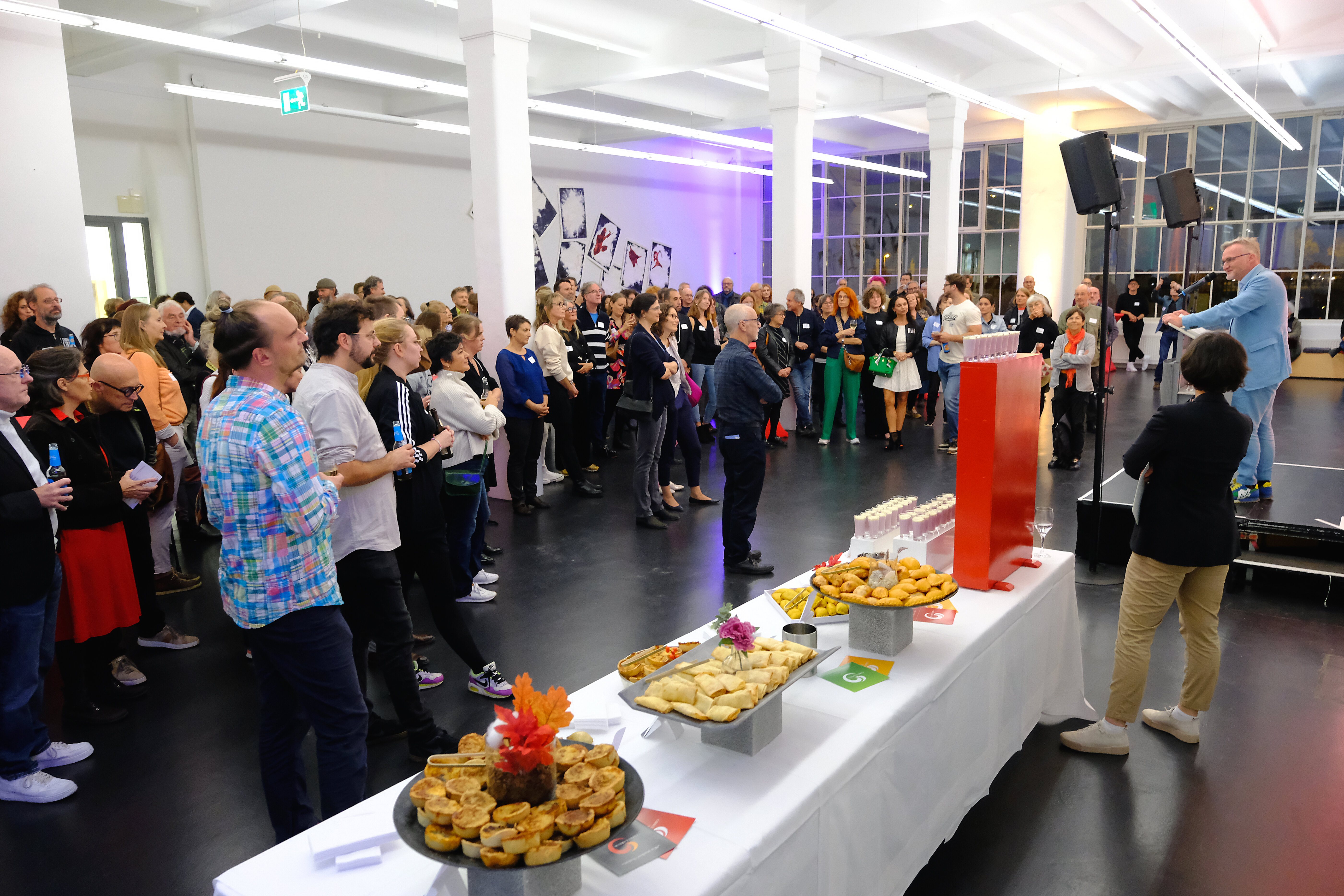 Viele Menschen in einem Saal. Ein Mann spricht ins Mikrofon. Ein Buffet steht bereit