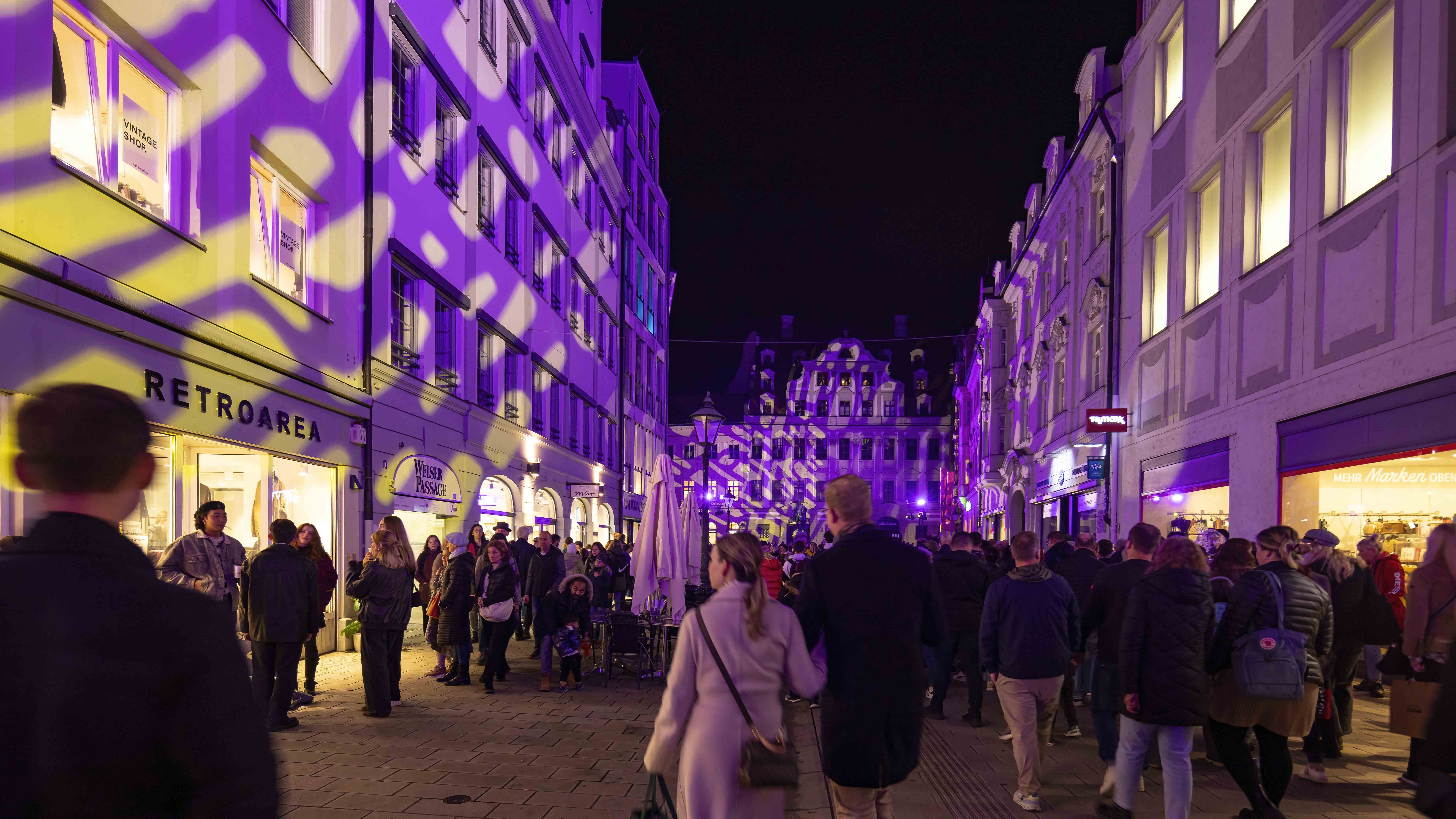 Auf dem Foto ist eine briete Straße mit vielen Geschäften zu sehen. Die Fassaden der Gebäude sind bunt angeleuchtet und es sind viele Menschen unterwegs