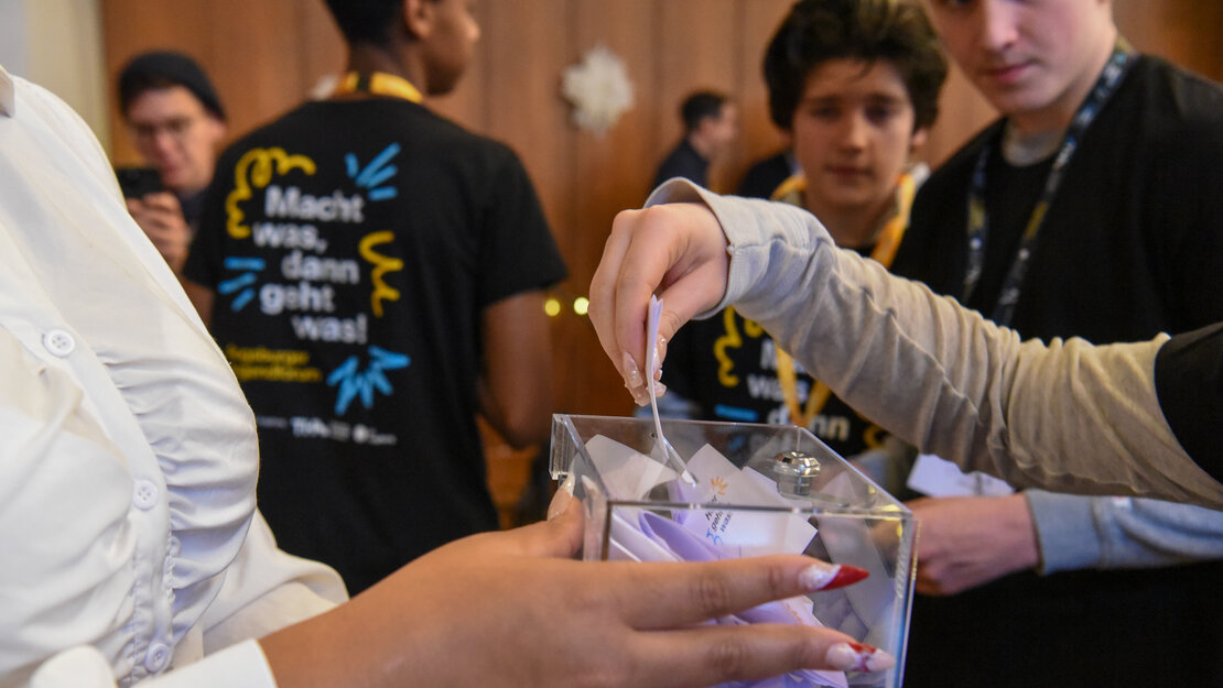 jemand hält eine Kunststoffbox, in den Schlitz steckt jemand anderer einen Zettel. Im Hintergrund steht jemand mit einem T-Shitrt, auf dem "Macht was, dann geht was" steht