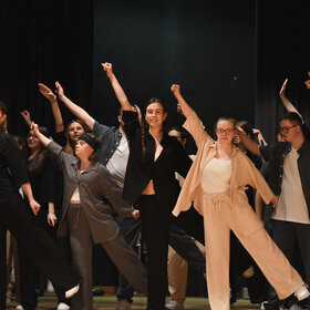 Leute, die überwiegend jung und dunkel gekleidet sind, tanzen: sie stehen auf einem Bein und haben einen Arm schräg in die Luft erhoben