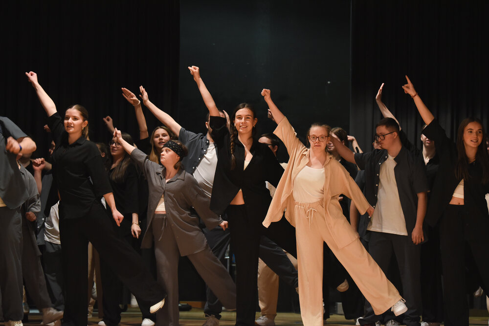 Leute, die überwiegend jung und dunkel gekleidet sind, tanzen: sie stehen auf einem Bein und haben einen Arm schräg in die Luft erhoben