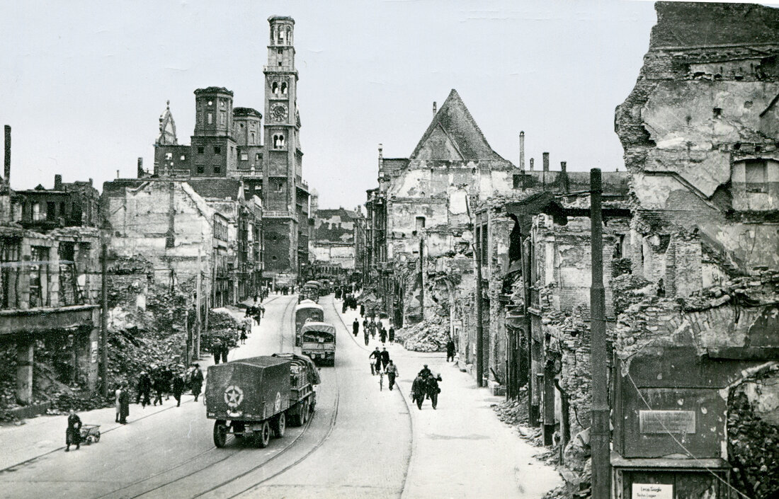 Das Schwarz-Weiß-Bild zeigt die Karolinenstraße mit Blick auf den Perlachturm im Jahr 1945: Die Häuser sind von Bomben zerstört. Auf der Straße fahren mehrere amerikanische Militärfahrzeuge. Fußgänger laufen am Straßenrand.