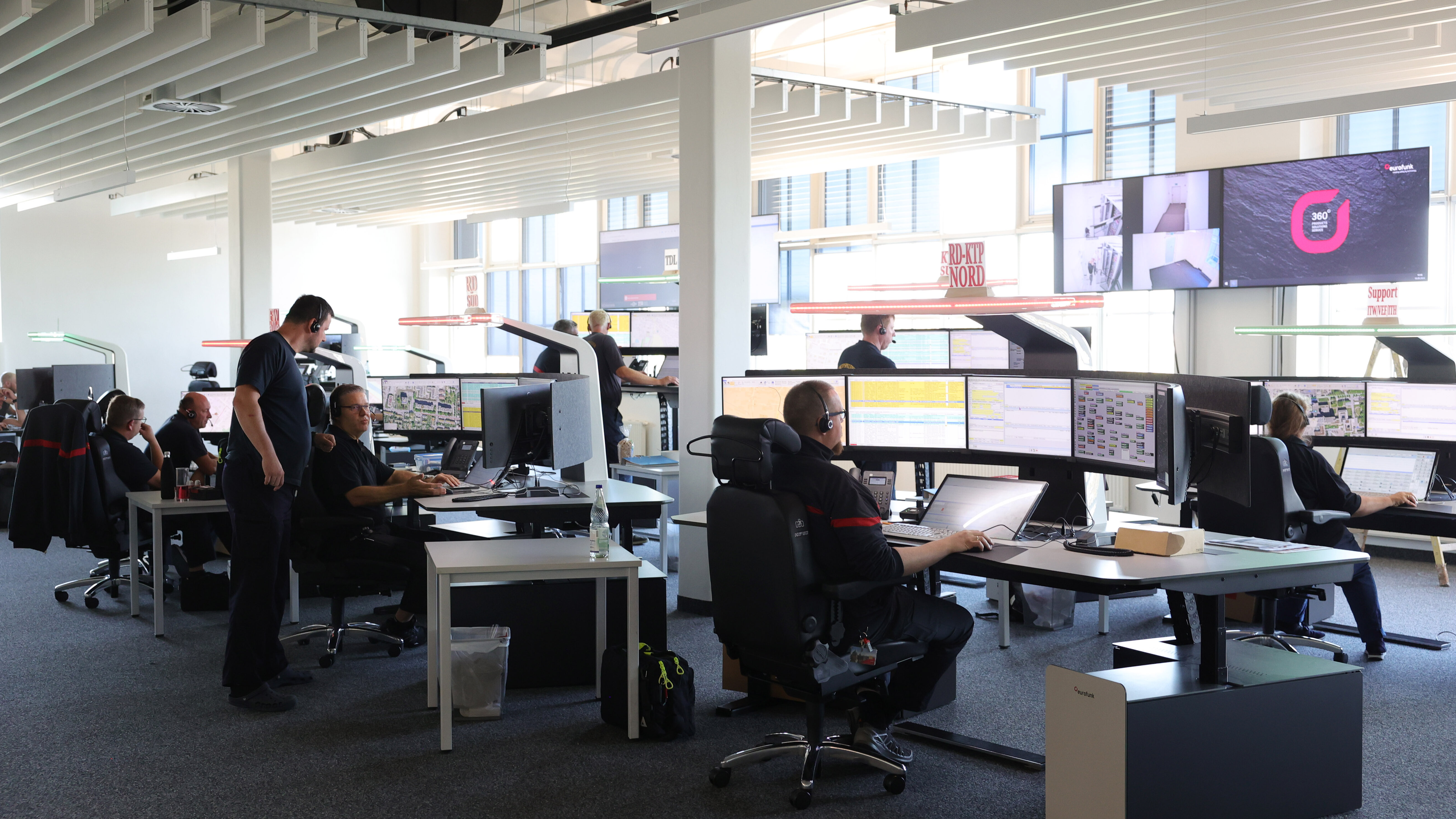 in einer großen Halle mit offenliegender Decke stehen viele Büromöbel und Computer, an der Wand große Bildschirme und Anzeigetafeln