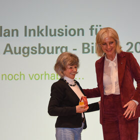 zwei Frauen lächeln und berühren sich, die eine ist groß und blond, die andere klein und hellhaarig. Sie trägt ein kleines Haus auf der Handfläche