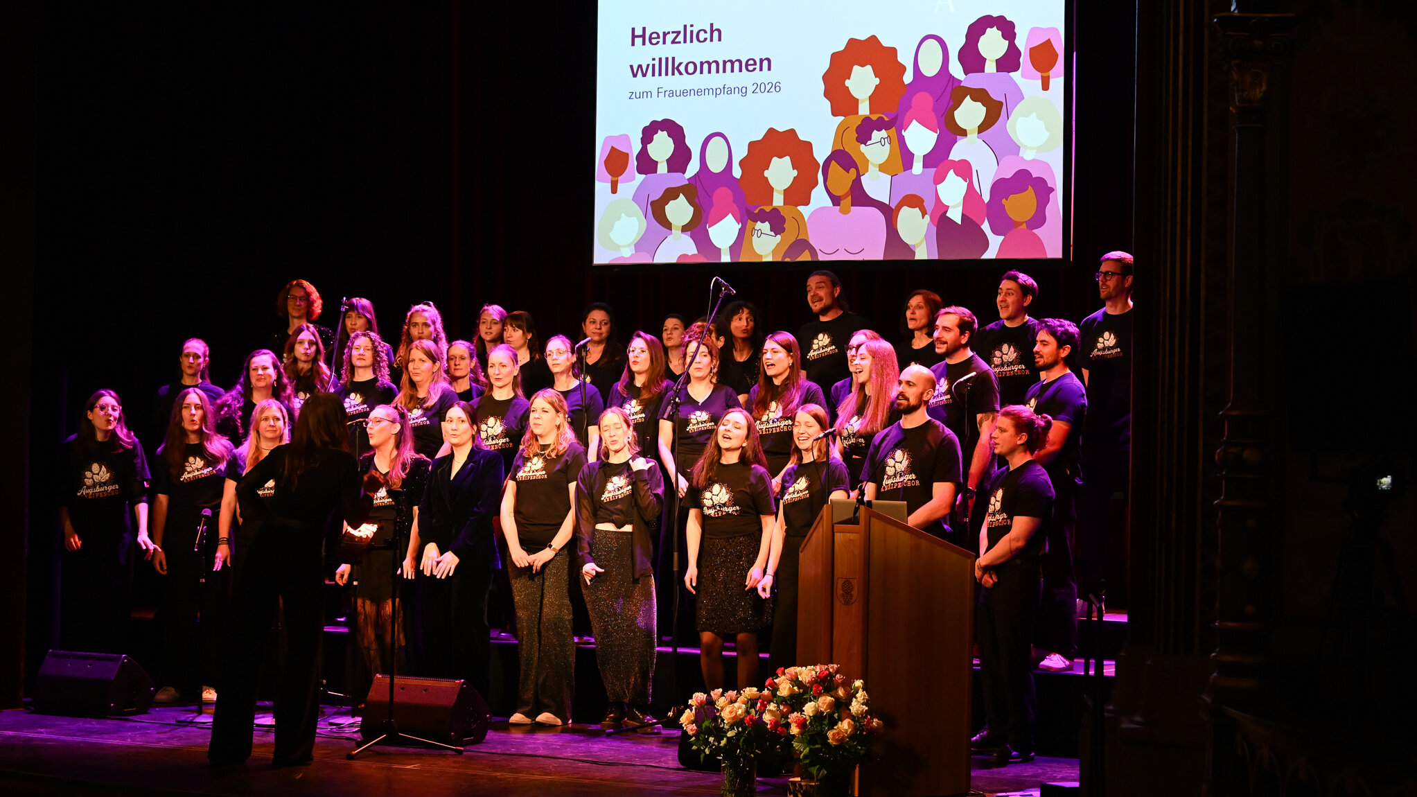 Das Foto zeigt viele Menschen, die in vier Reihen auf einer Bühne stehen. Alle tragen schwarze T-Shirts mit der Aufschrift Augsburger Kneipenchor
