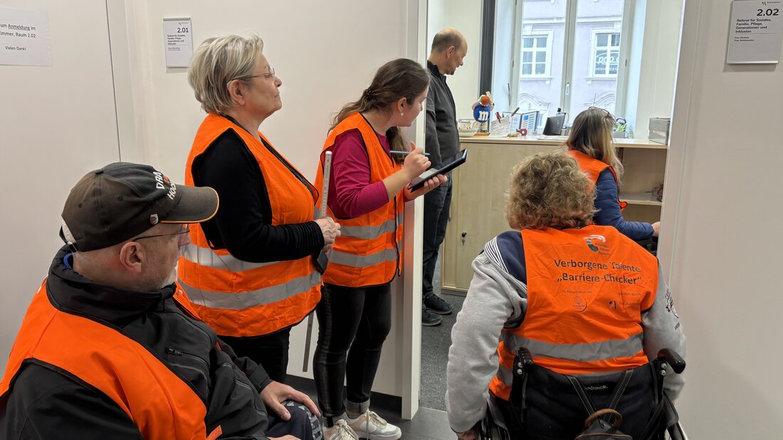Fünf Barriere-Checkerinnen und -Checker in neonorangenen Warnwesten gehen in den Räumlichkeiten des Sozialreferats einen Fragebogen zur Barrierefreiheit durch. Eine Rollstuhlfahrerin sieht sich dazu im Geschäftszimmer um, während die anderen aus dem Flur die Breite der Türe begutachten.