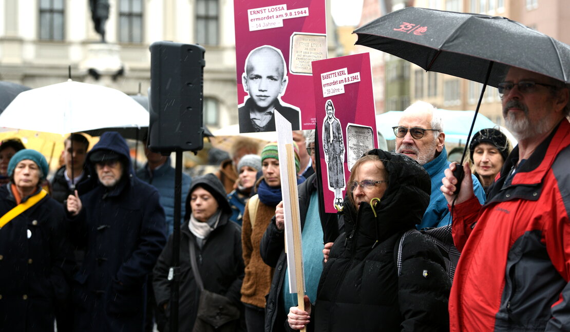 Menschen mit Regenschirmen stehen auf dem Rathausplatz, einige halten Schilder mit Bildern von Augsburger Holocaust-Opfern in die Höhe