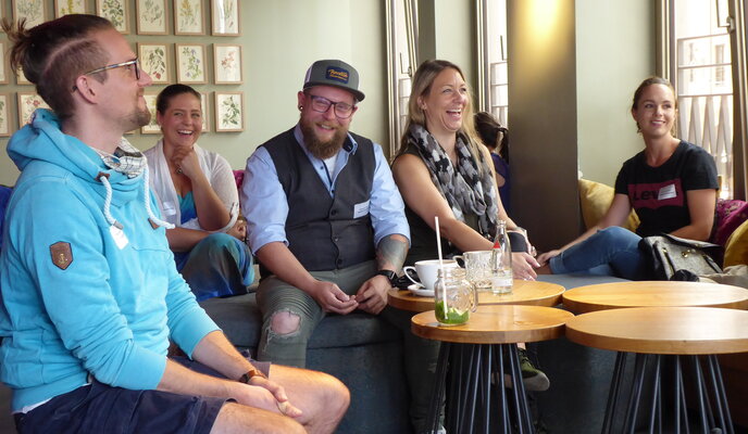 5 Menschen sitzen in einem Café an kleinen Tischen und lachen
