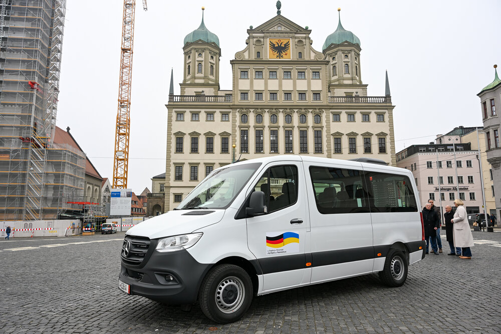 Das Foto zeigt einen weißen Kleinbus auf dem Rathausplatz. An der Fahrertür ist eine deutsche und eine ukrainische Flagge abgebildet