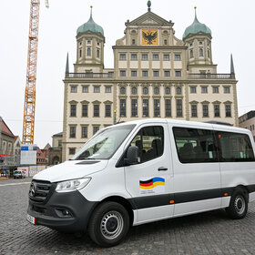 Das Foto zeigt einen weißen Kleinbus auf dem Rathausplatz. An der Fahrertür ist eine deutsche und eine ukrainische Flagge abgebildet
