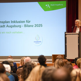 eine andere blonde Frau mit Brille am Rednerpult, neben ihr eine Leinwand, auf die "Aktionsplan Inklusion der Stadt Augsburg - Bilanz 2025" projiziert ist