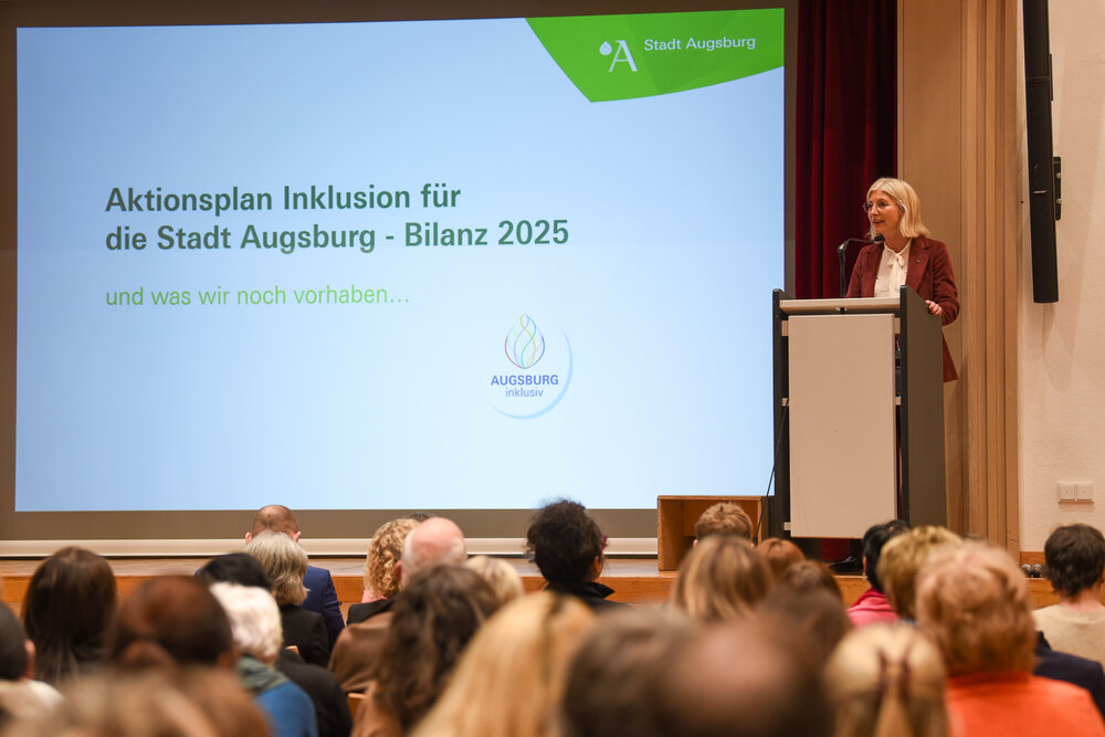 eine andere blonde Frau mit Brille am Rednerpult, neben ihr eine Leinwand, auf die "Aktionsplan Inklusion der Stadt Augsburg - Bilanz 2025" projiziert ist