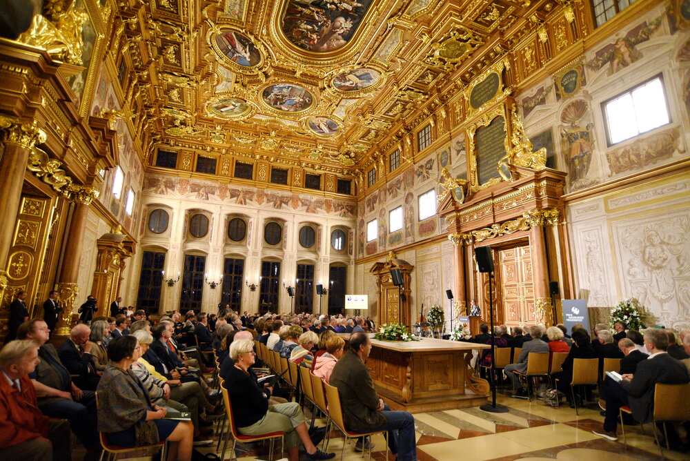 In einem großen Saal sitzen viele Menschen auf Stühlen. Im Bildhintergrund hält eine Frau eine Rede.