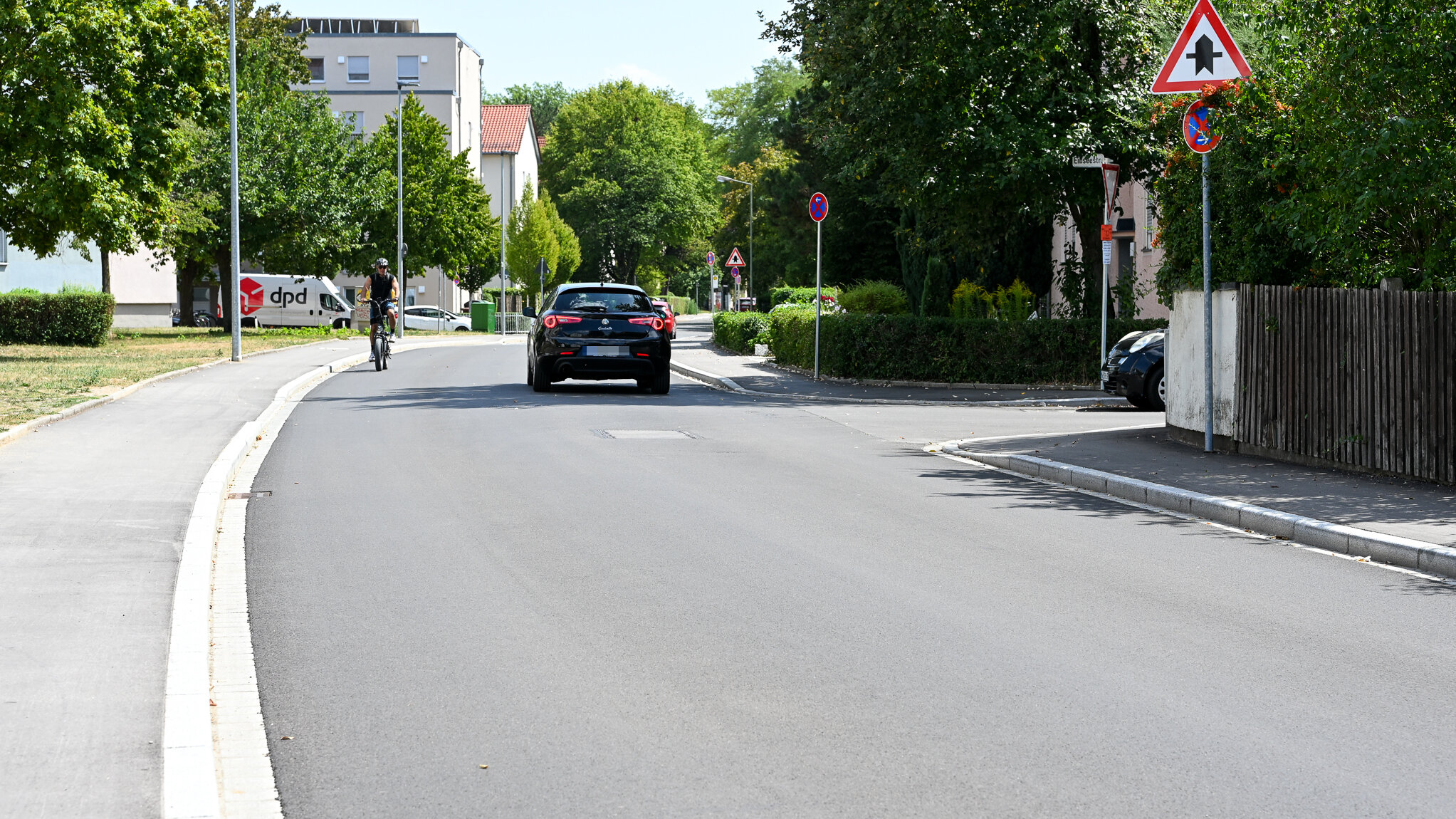 eine Straße mit neuer Teerdecke, rechts und links Gebäude und Vorgärten mit Bäumen und Büschen