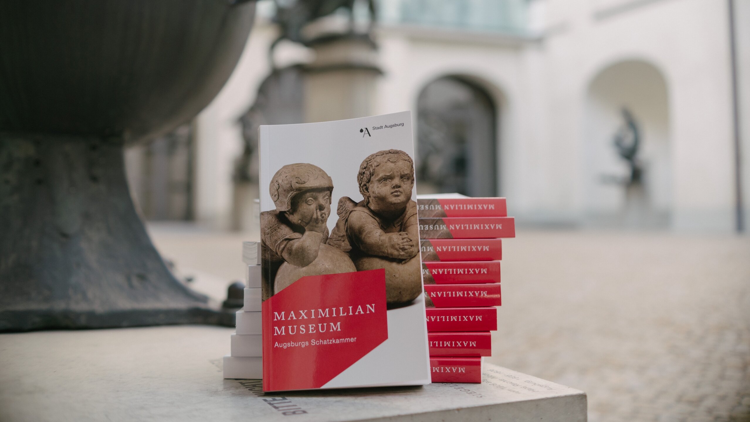 Im Vordergrund ist ein Buch mit der Überschrift Maximilianmuseum Augsburgs Schatzkammer zu sehen. Im Hintergrund ist ein gepflasterter Hof mit Skulpturen zu sehen.