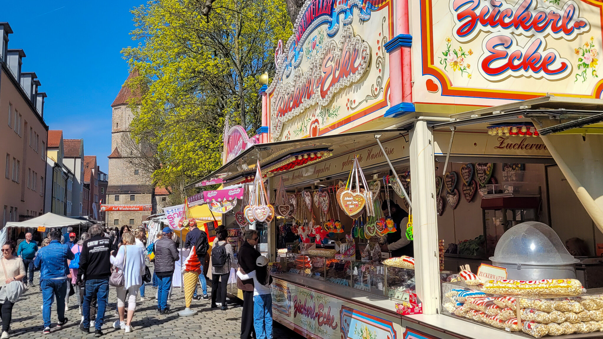 Blick in die Dult. Viele Leute spazieren zwischen den Ständen. Im Vordergrund ein Süßwaren-Stand, im Hintergrund das Jakobertor.