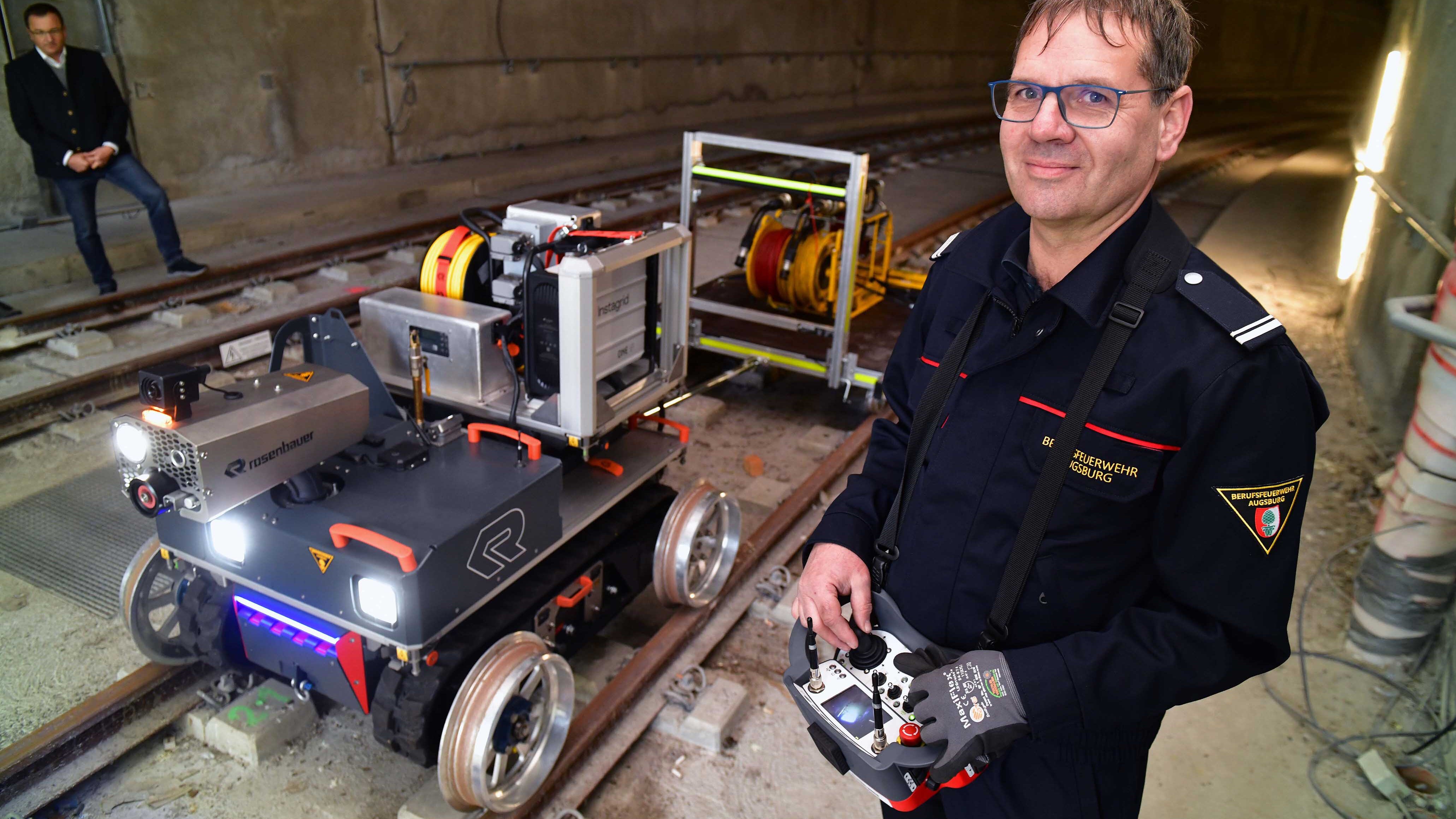 Ein Mann in Feuerwehr-Uniform hält eine Fernbedienung in der Hand. Hinter ihm steht ein kleines Gefährt auf Gleisen