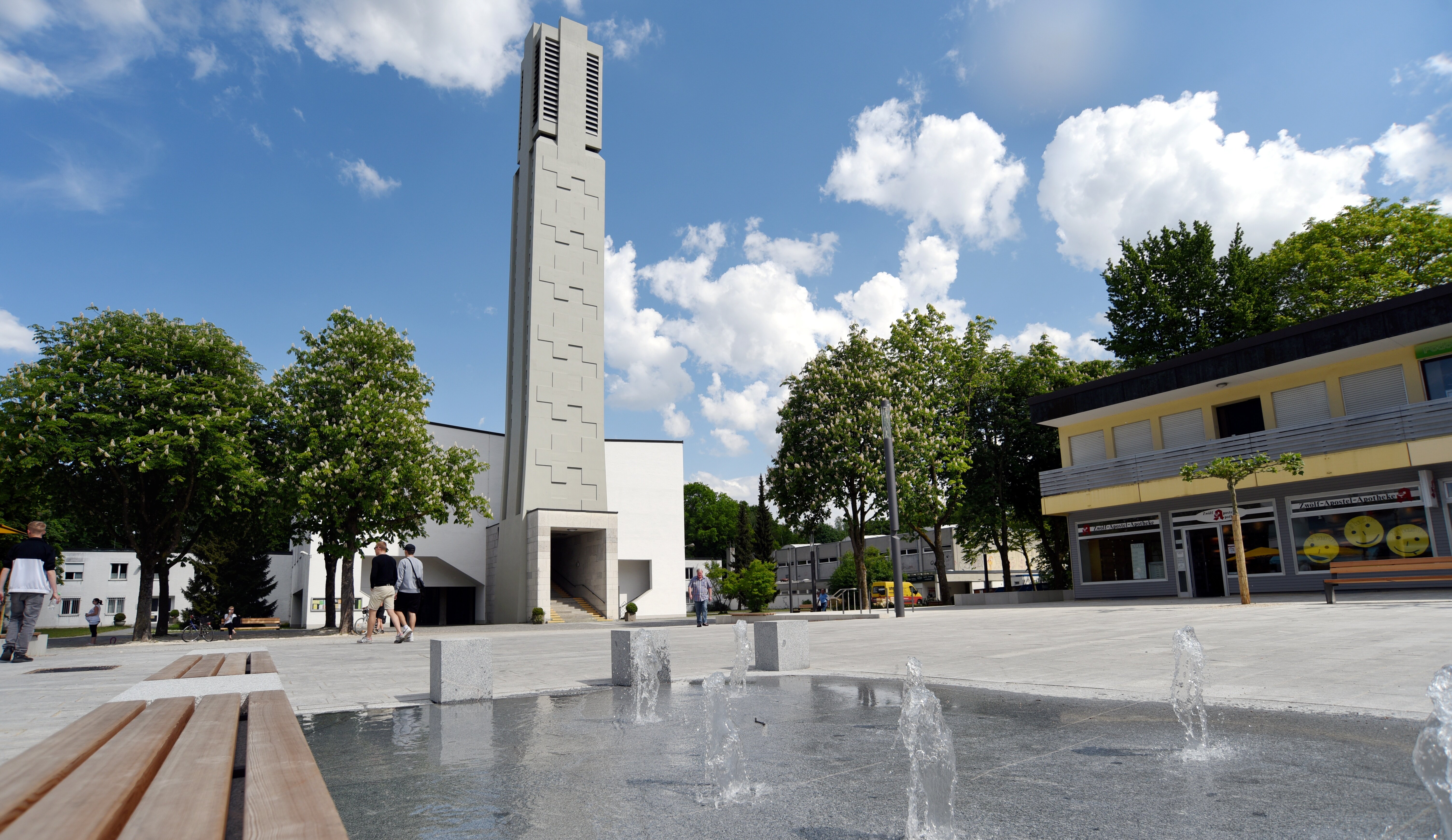 Ein Platz mit einem Brunnen und Fontänen, Geschäften und einem modernen Kirchturm