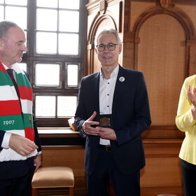 Drei Personen stehen im Rathaus. Der Mann in der Mitte hält eine Medaille. Links ein Mann mit einem FCA-Schal. Rechts eine Frau. Sie applaudiert.