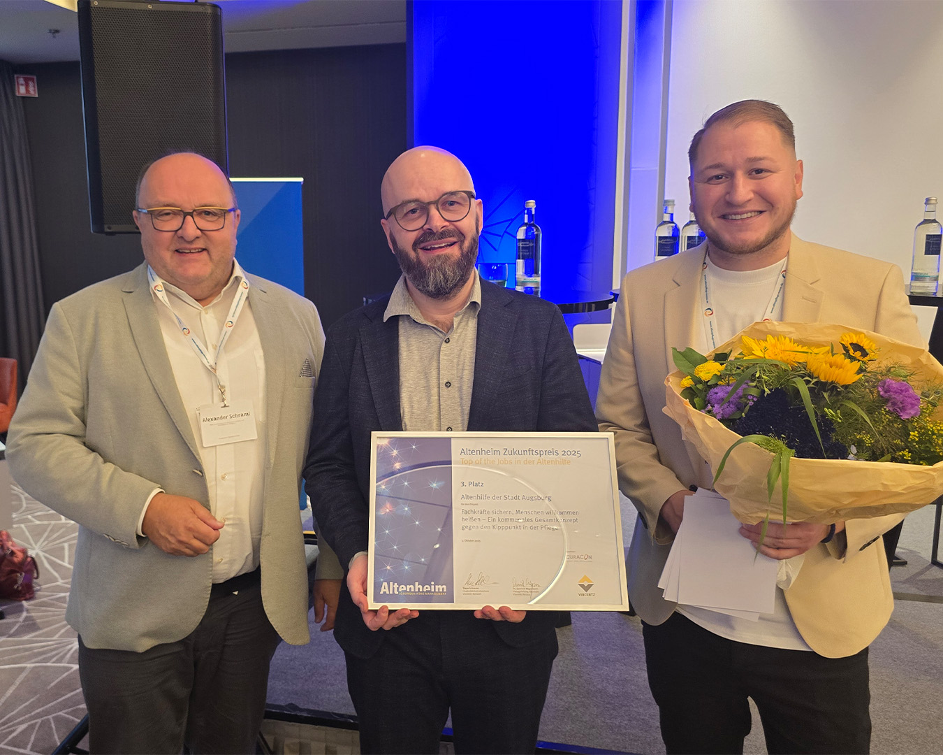 Drei Männer halten eine Urkunde und einen Blumenstrauß