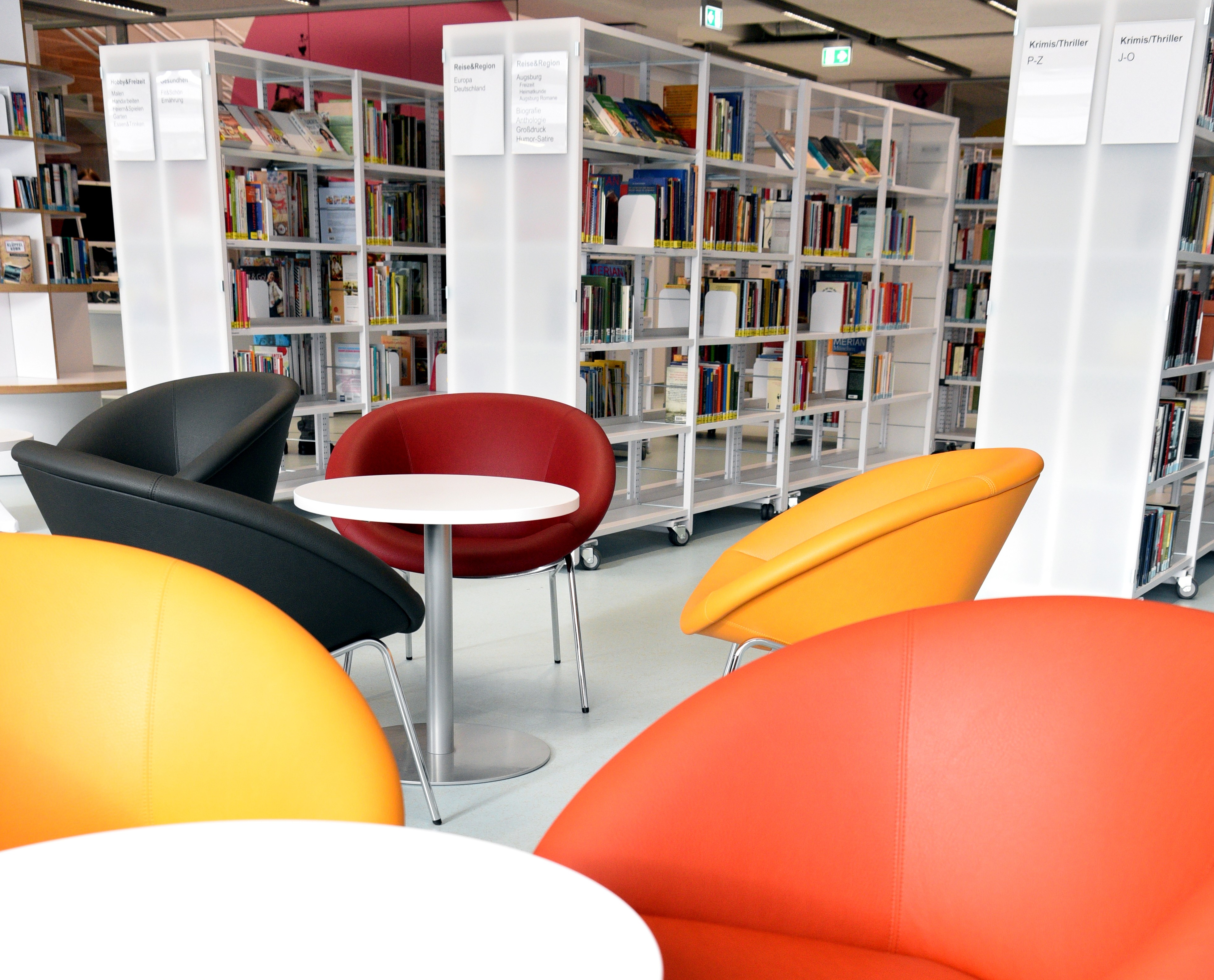 Die Stadtteilbücherei Lechhausen von innen: bunte Stühle stehen an einem Tisch im Vordergrund. Hinten sind mehrere Bücherregale zu sehen.