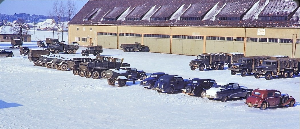 Im Hintergrund ein lang gezogenes gelbes Gebäude, offenbar eine Lagerhalle. Es liegt Schnee. Vor der Halle stehen zahlreiche Fahrzeuge aus den 1950er Jahren.