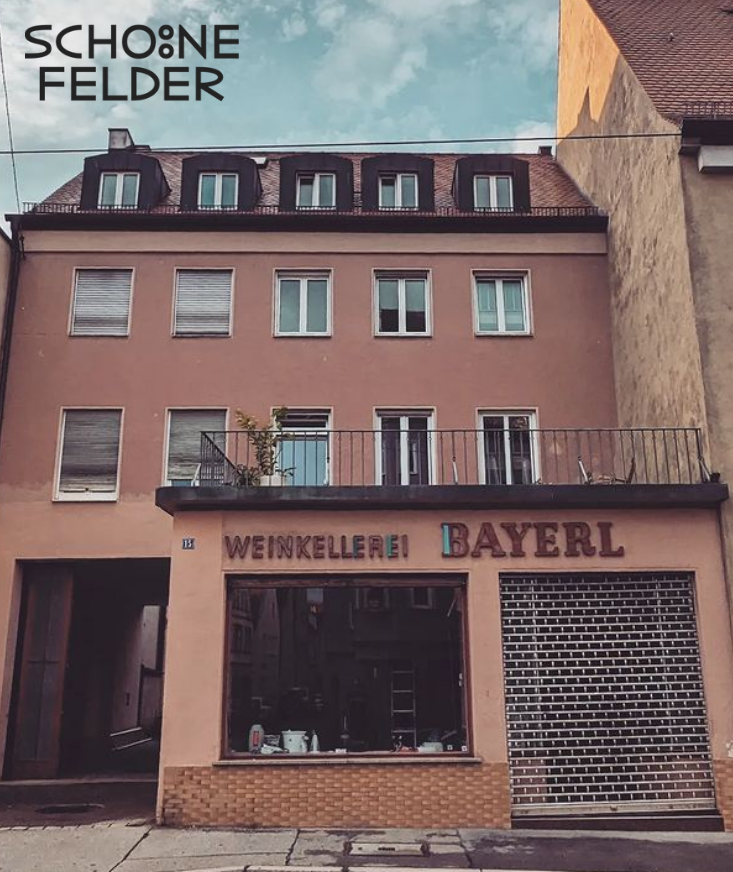 Ein rotes Haus mit der Aufschrift "Weinkellerei Bayerl" ist zu sehen. Auf dem Foto ist die Aufschrift "Schöne Felder"