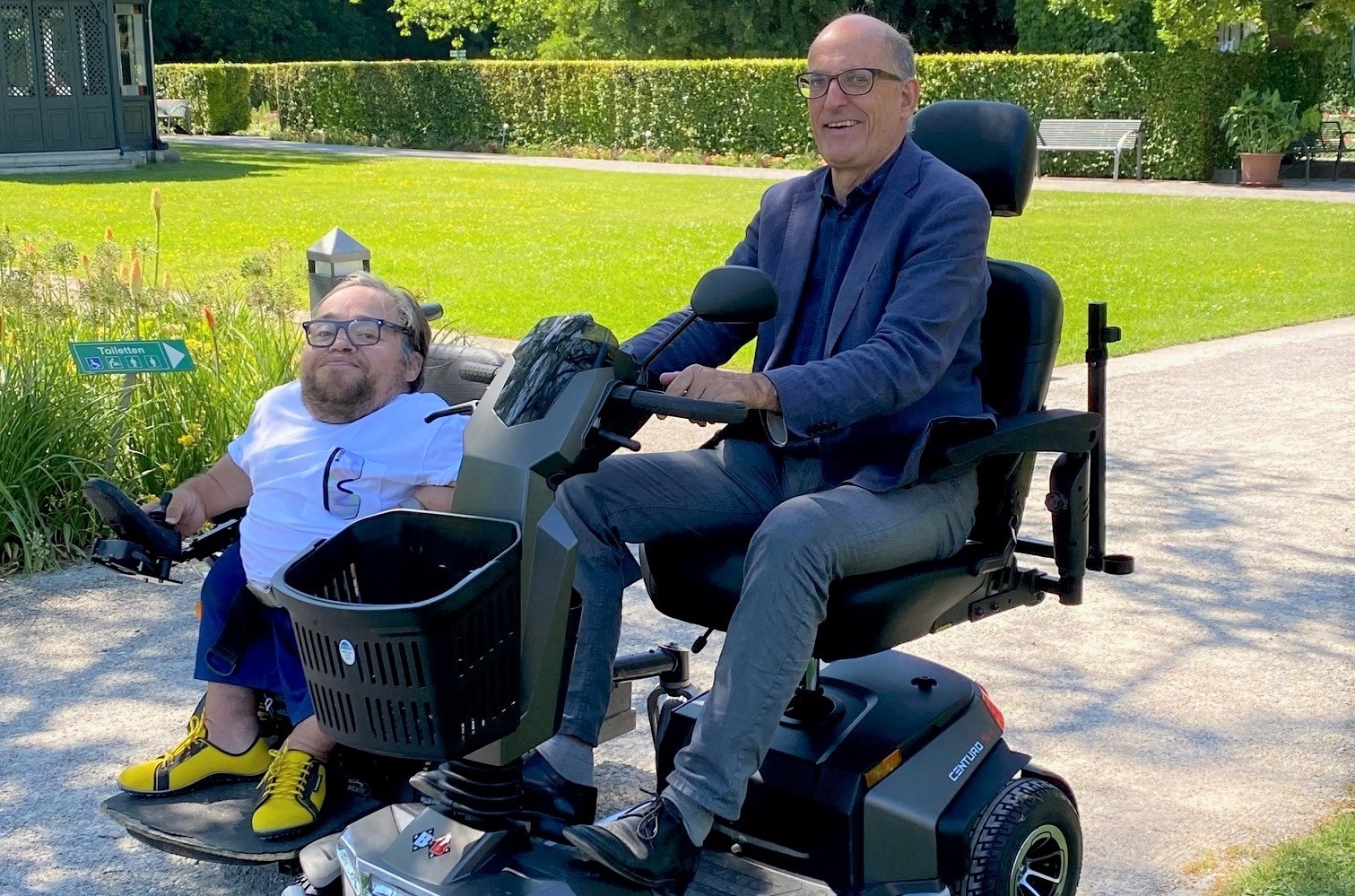 Ein Mann sitzt in einem Elektromobil, ein weiterer Mann in einem Rollstuhl daneben