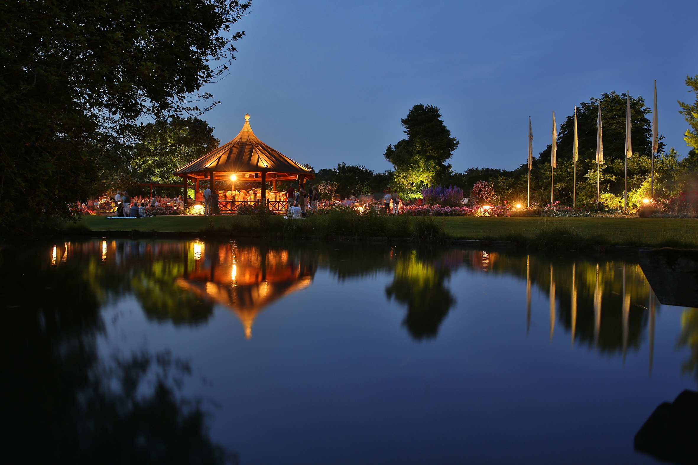Ein Pavillon im Botanischen Garten ist beleuchtet. Es ist Abend, der Himmel ist dunkelblau. Der Pavillon und die umliegenden Bäume spiegeln sich in einem kleinen See