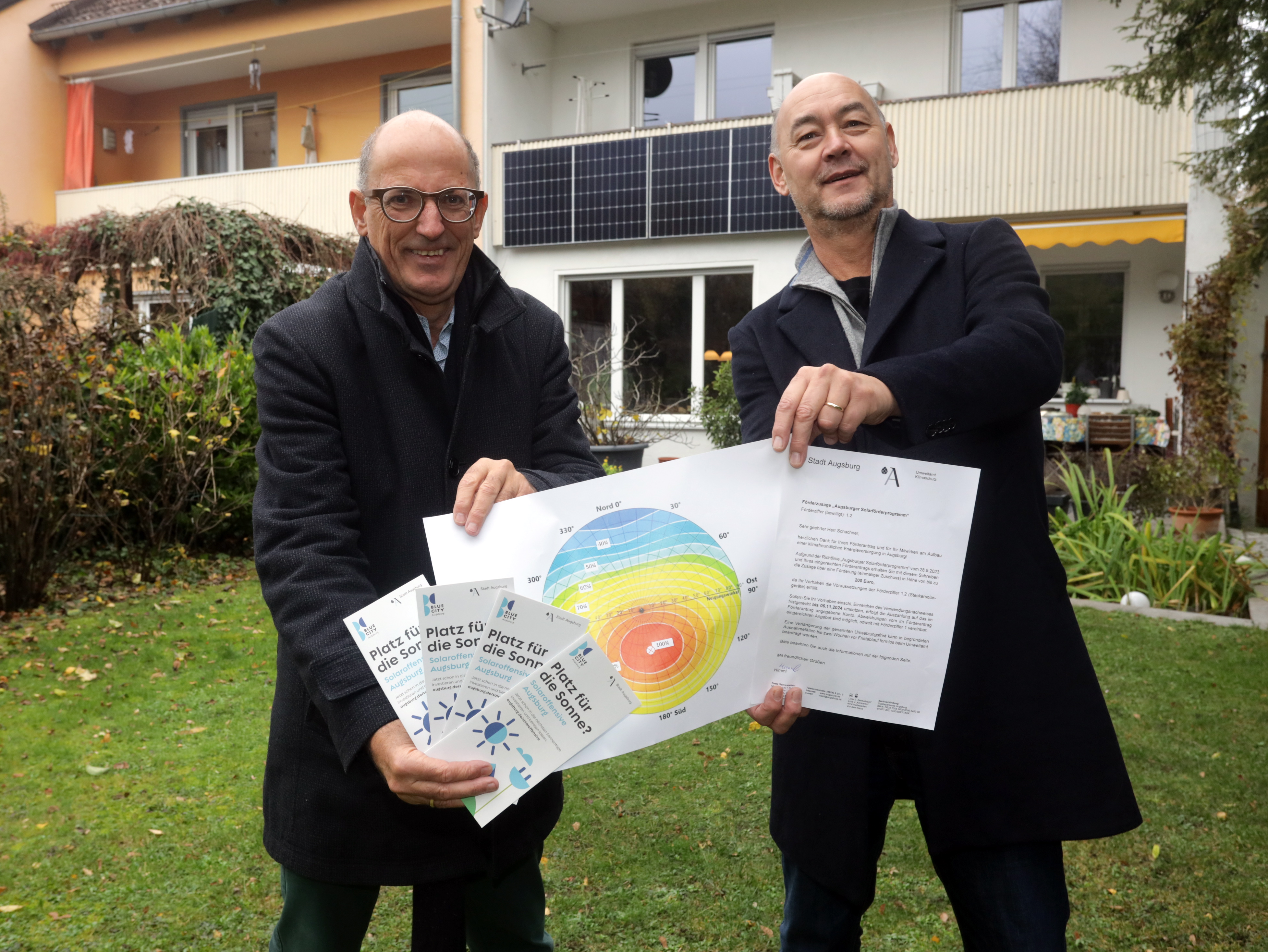 Zwei Männer stehen vor einem Reihenhaus im Garten. Sie halten viele Papiere in der Hand. Am Balkon im Hintergrund sind mehrere Solar-Module zu sehen.