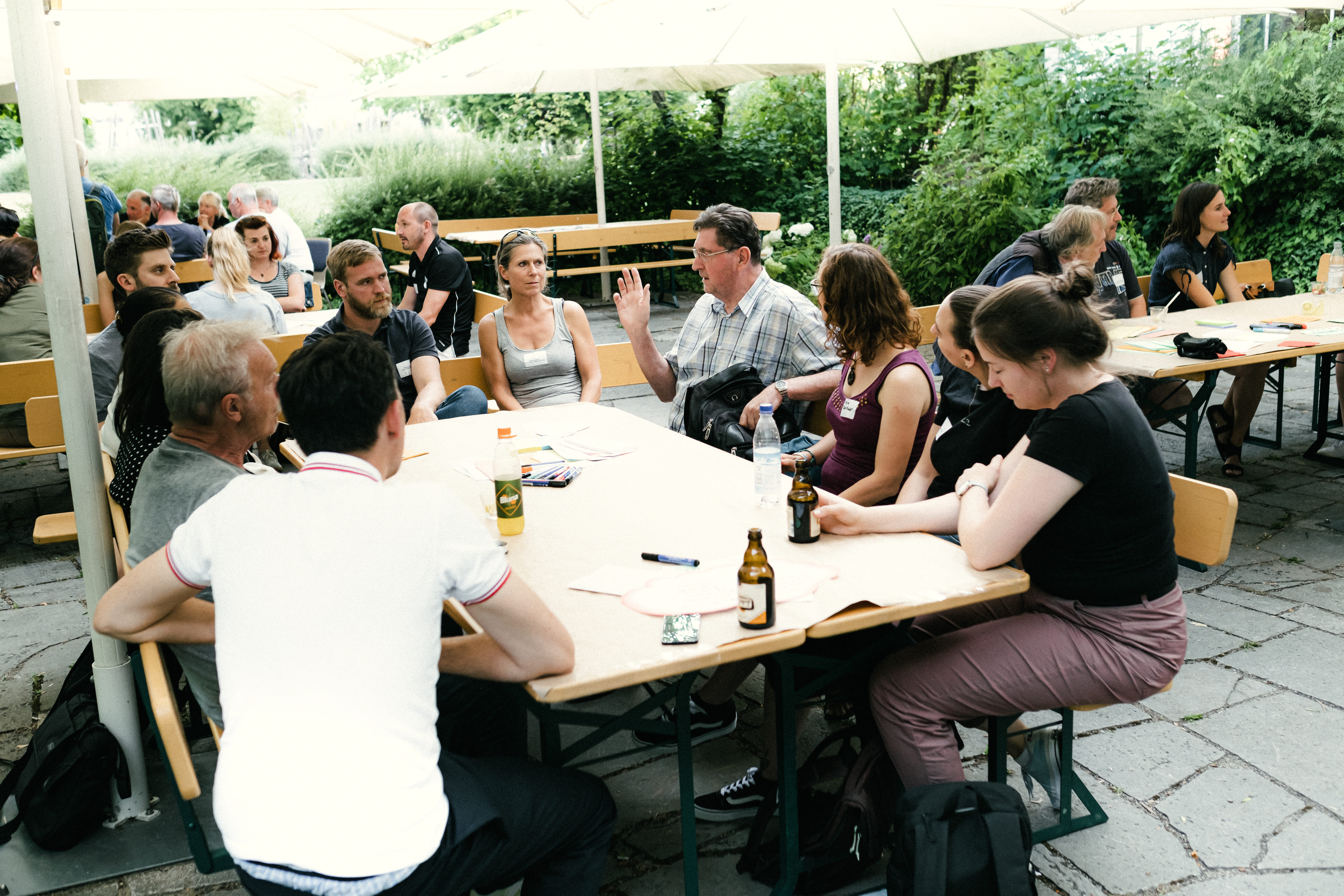 Etwa 25 Menschen sitzen in einem Biergarten an mehreren Tischen und unterhalten sich.