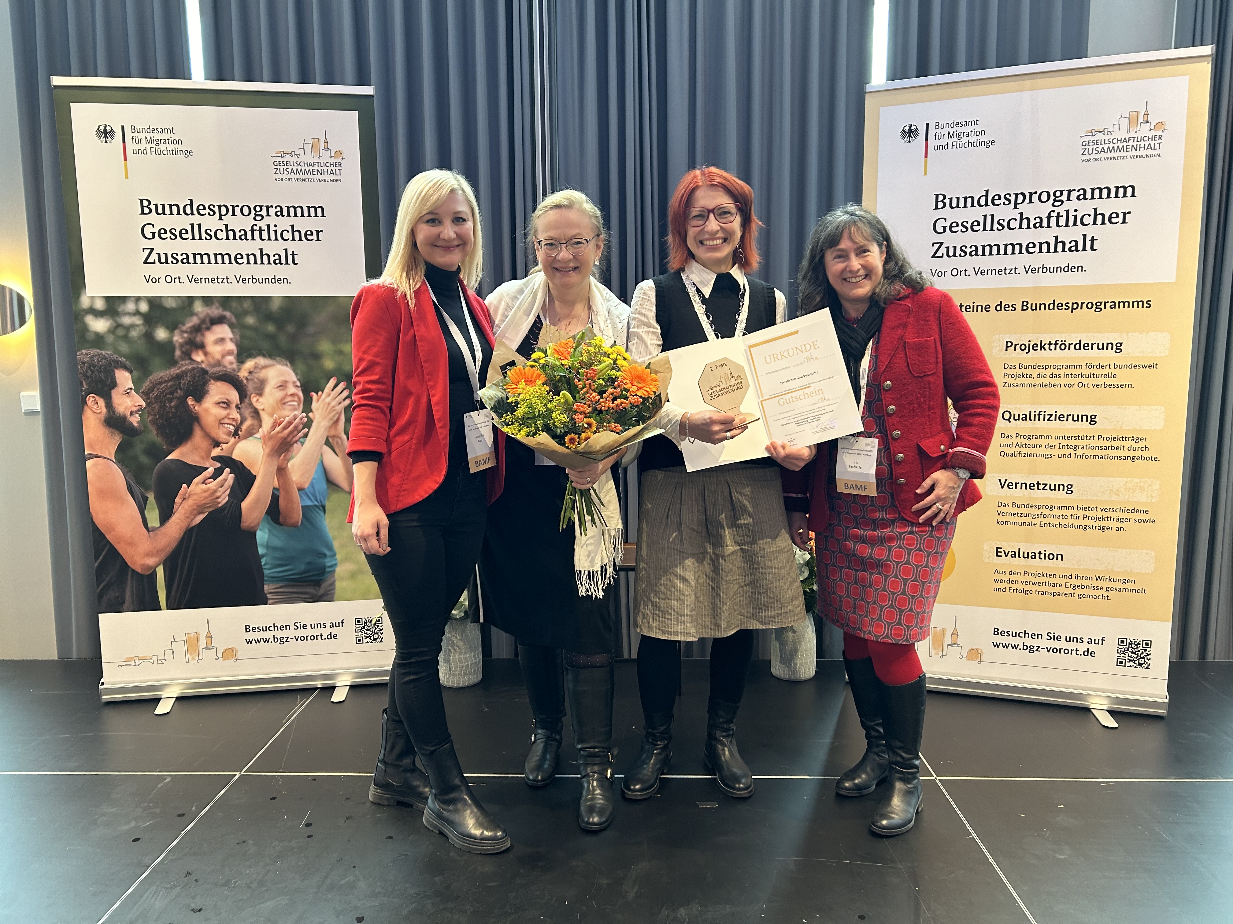 Vier Frauen stehen nebeneinander und lächeln in die Kamera. Eine von ihnen hält einen Blumenstrauß, eine andere eine Urkunde. Flankiert wird die Gruppe von zwei Roll-ups, auf denen "Bundesprogramm für gesellschaftlichen Zusammenhalt" steht.