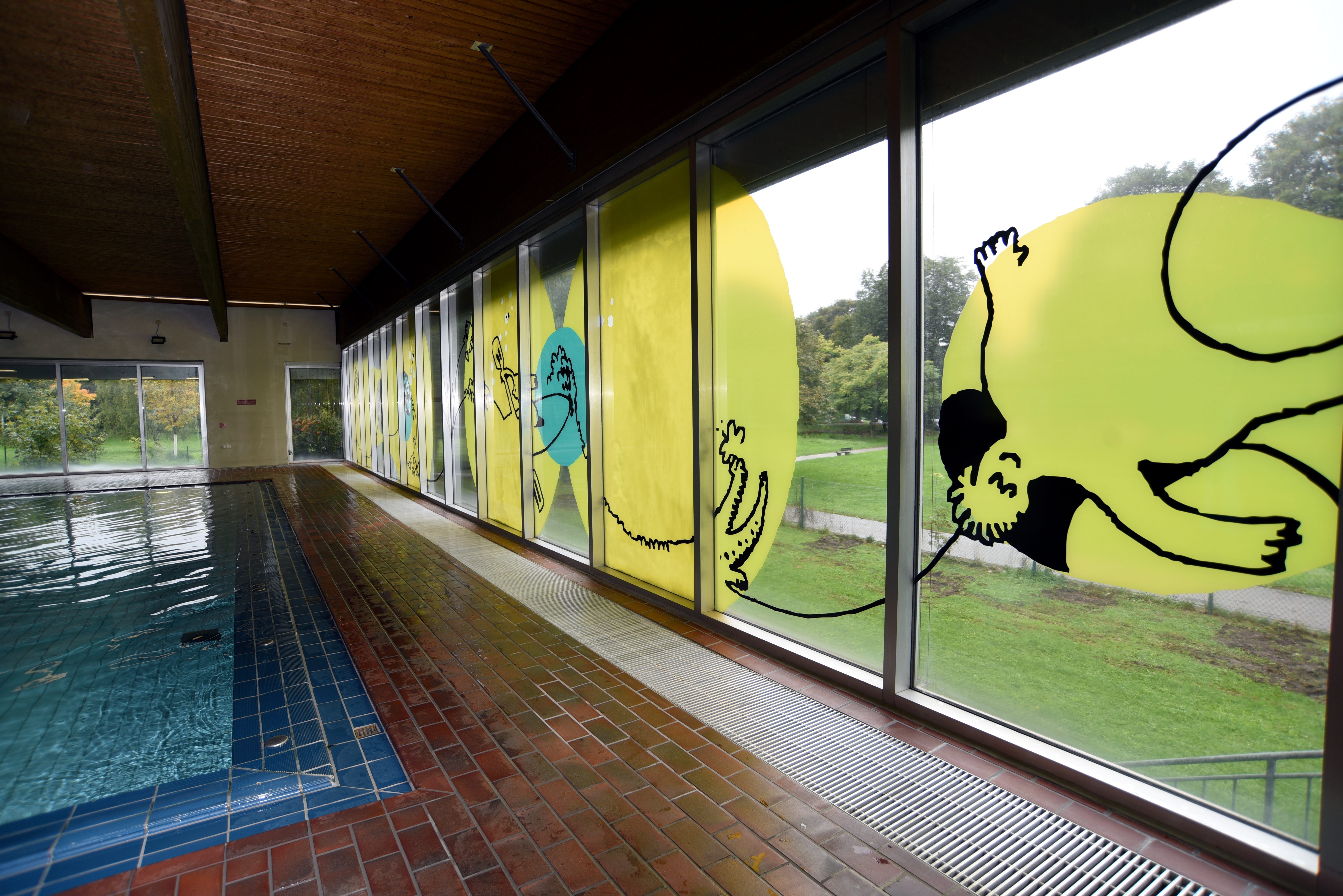 Die Fensterfront eines Hallenbades ist mit flächigen grafischen Elementen in gelb, blau und schwarz beklebt.